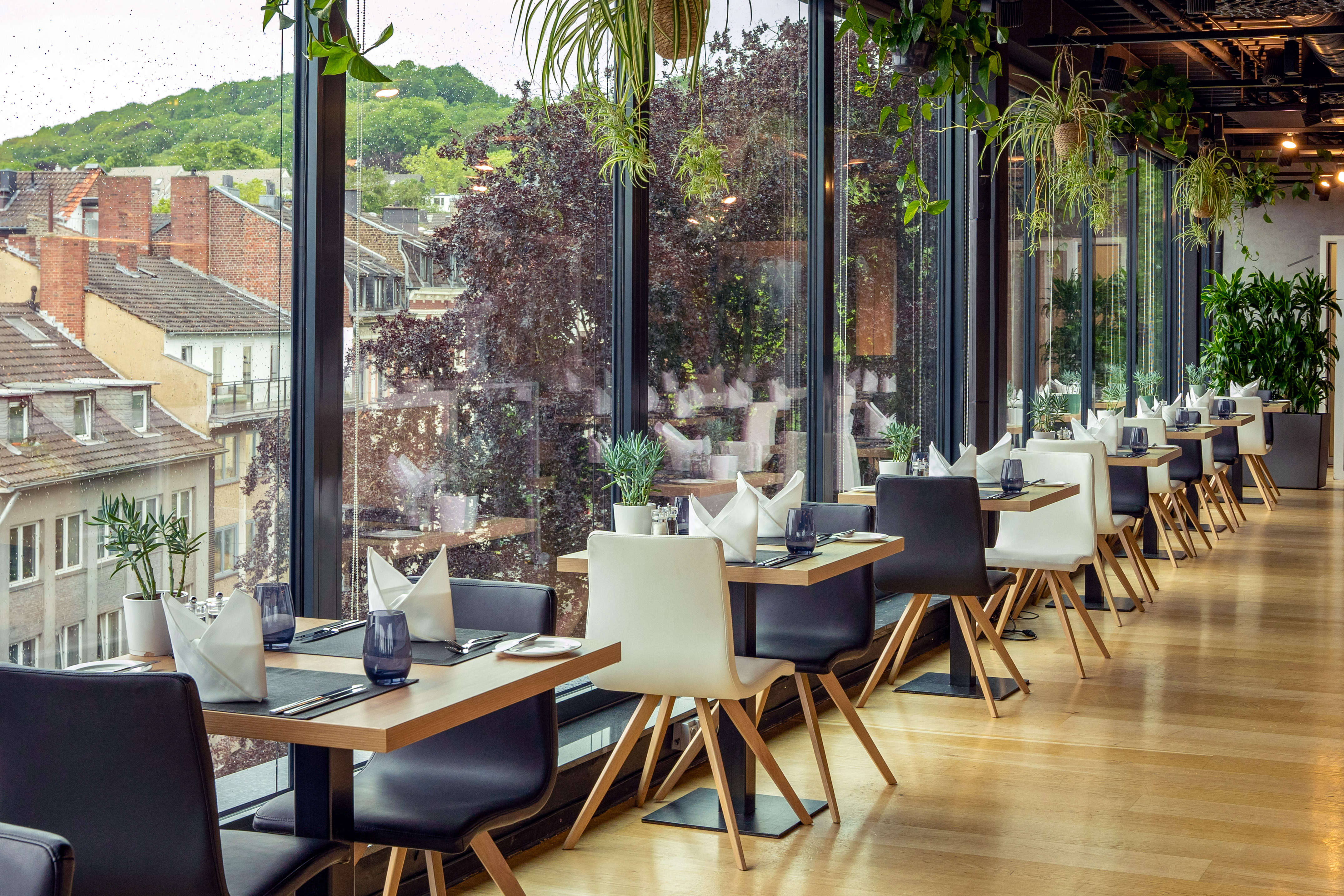 Modern restaurant with city view, large windows, set tables, and green plants.