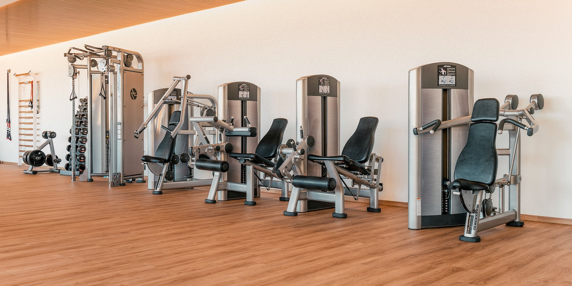 Modern gym with rows of strength training machines, dumbbells, and wooden floor.