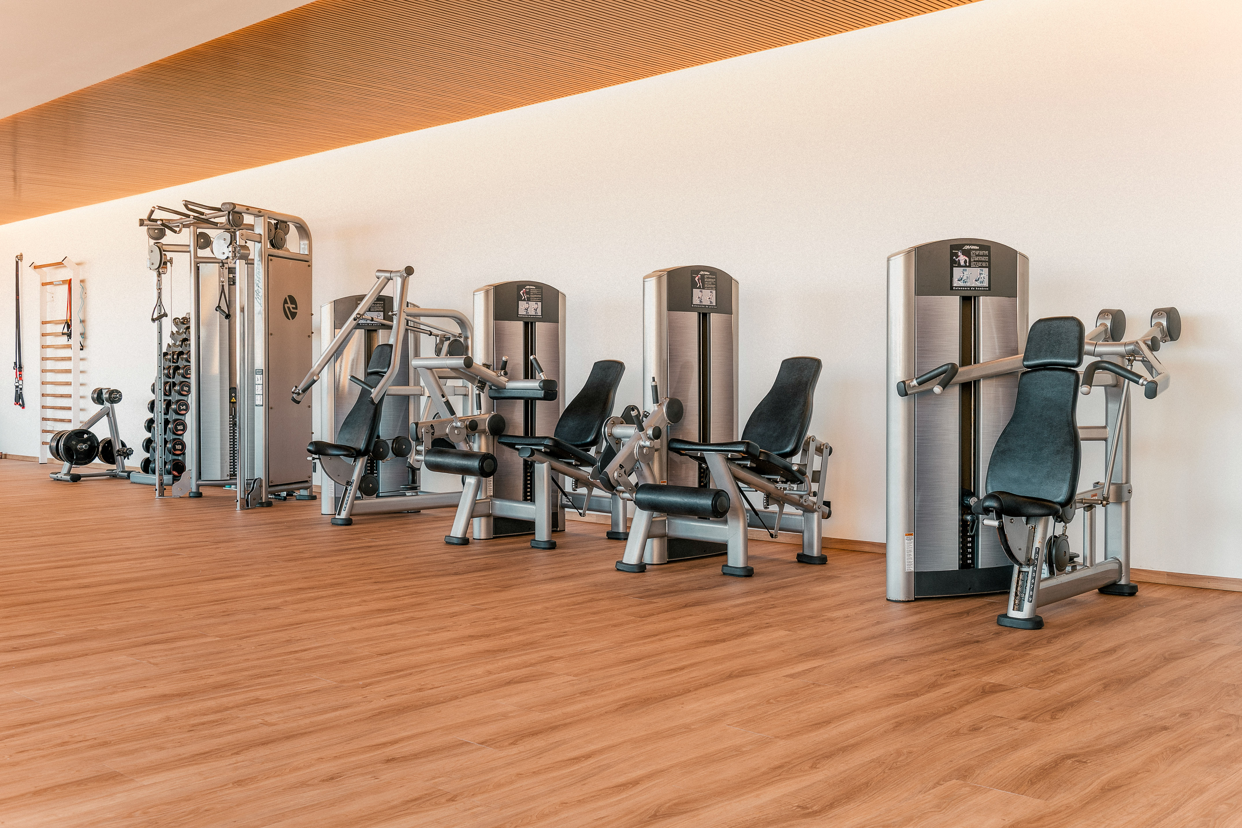 Modern gym with rows of strength training machines, dumbbells, and wooden floor.