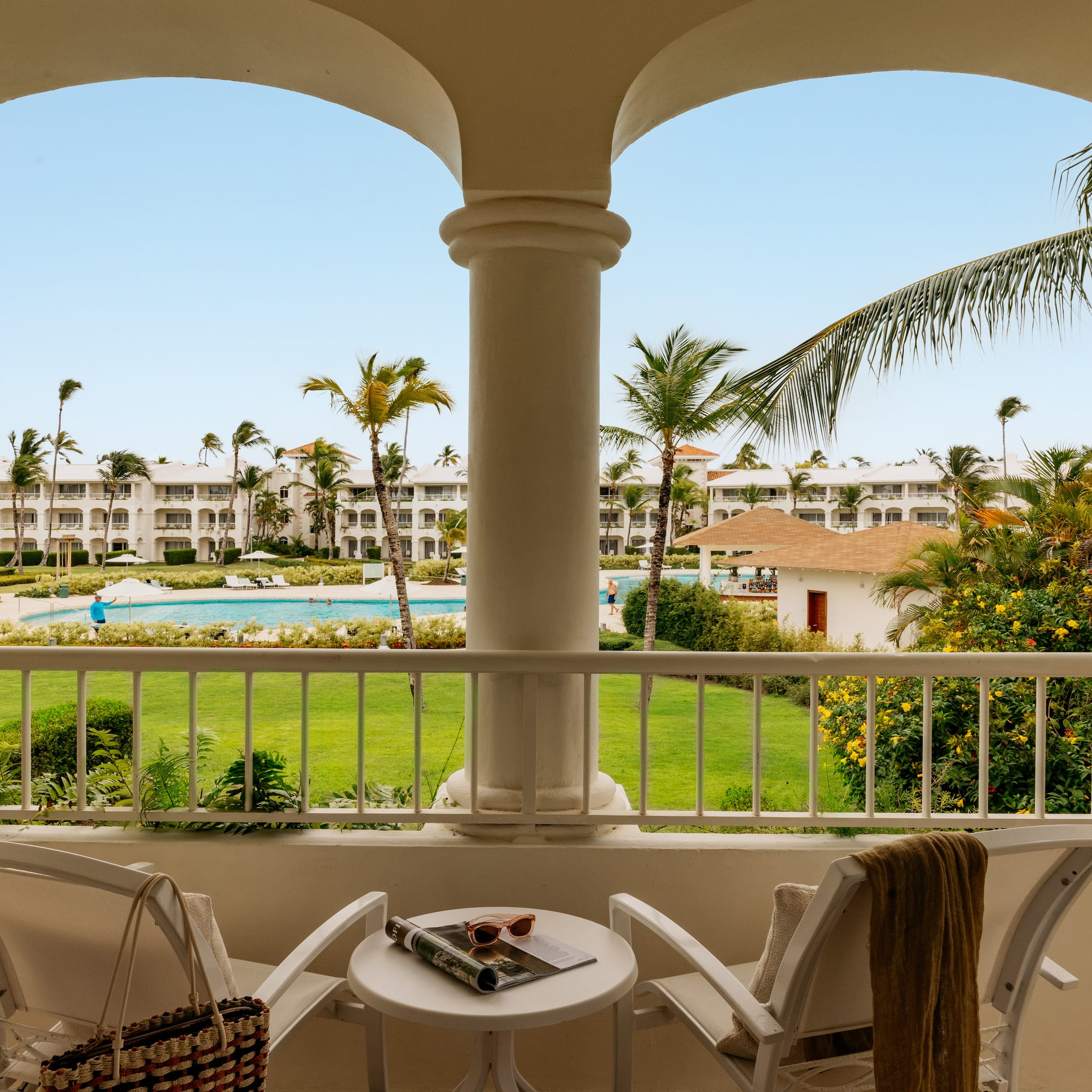 Tropical resort balcony overlooking pool, lush gardens, and white buildings.