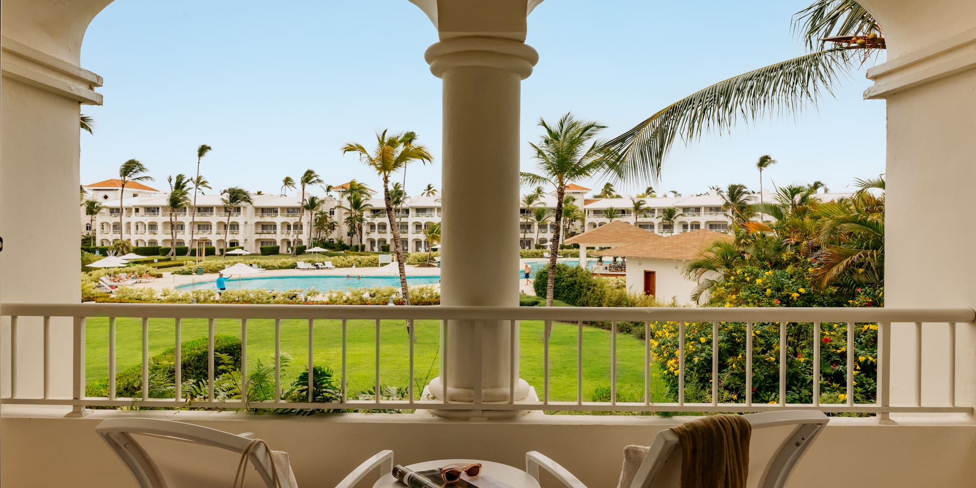Tropical resort balcony overlooking pool, lush gardens, and white buildings.