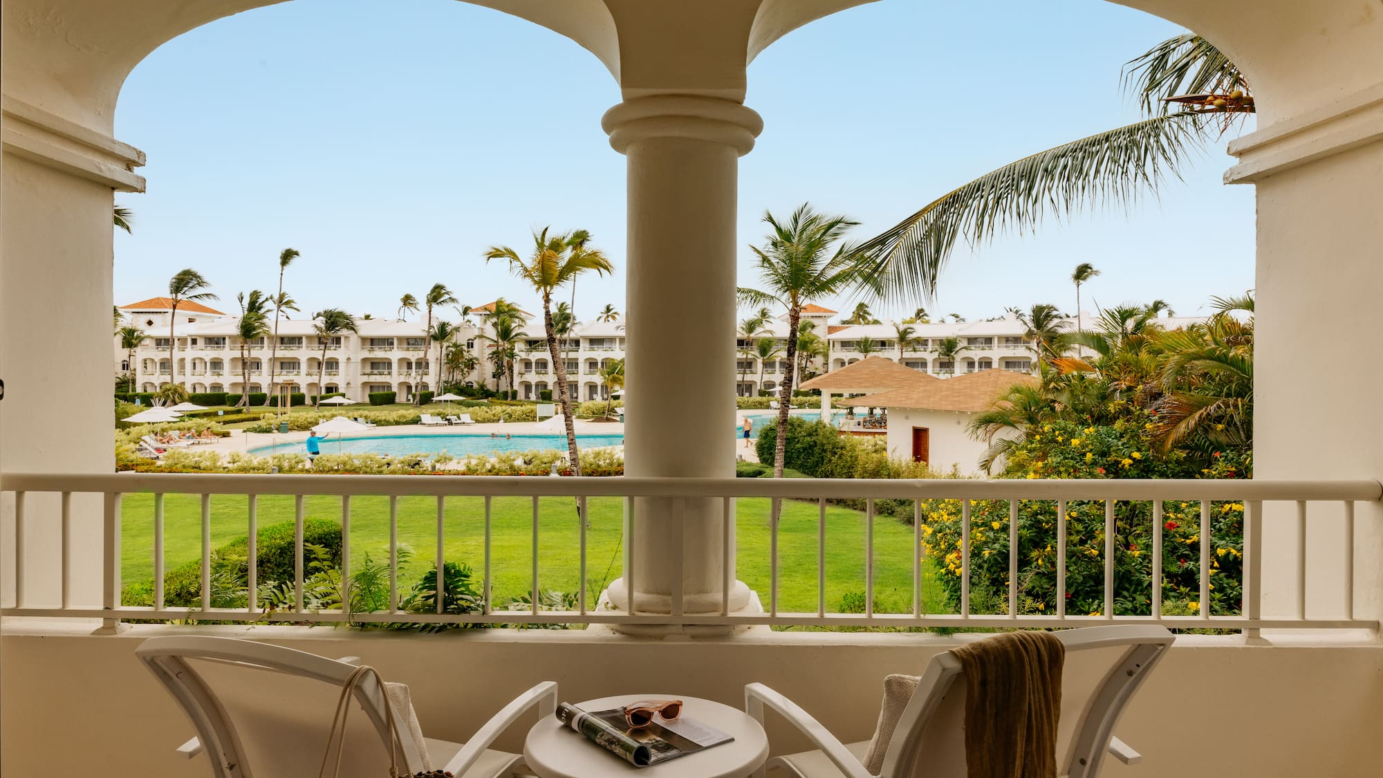 Tropical resort balcony overlooking pool, lush gardens, and white buildings.
