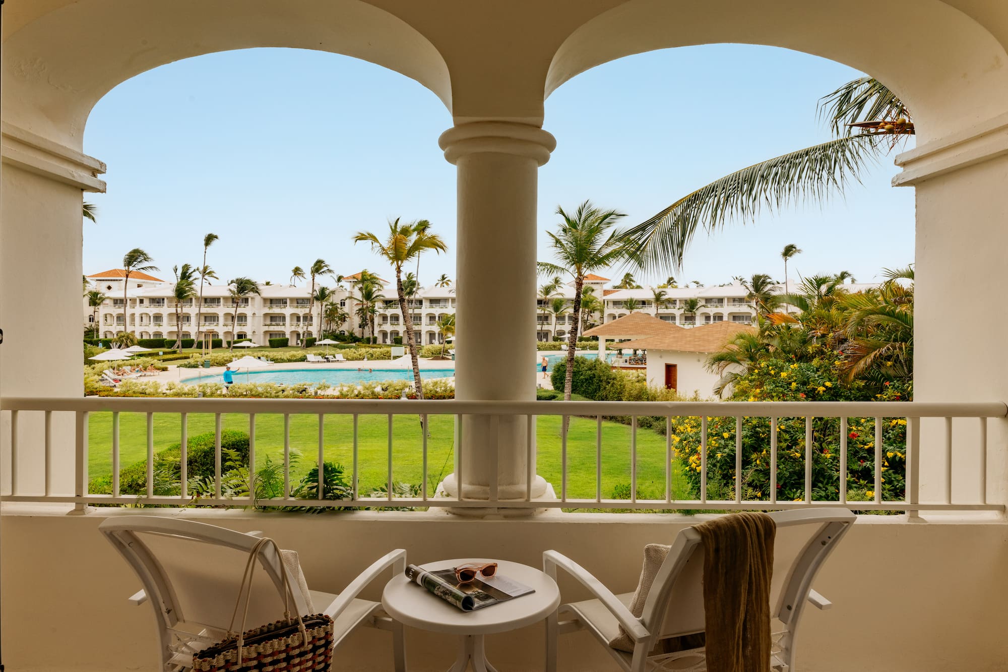 Tropical resort balcony overlooking pool, lush gardens, and white buildings.