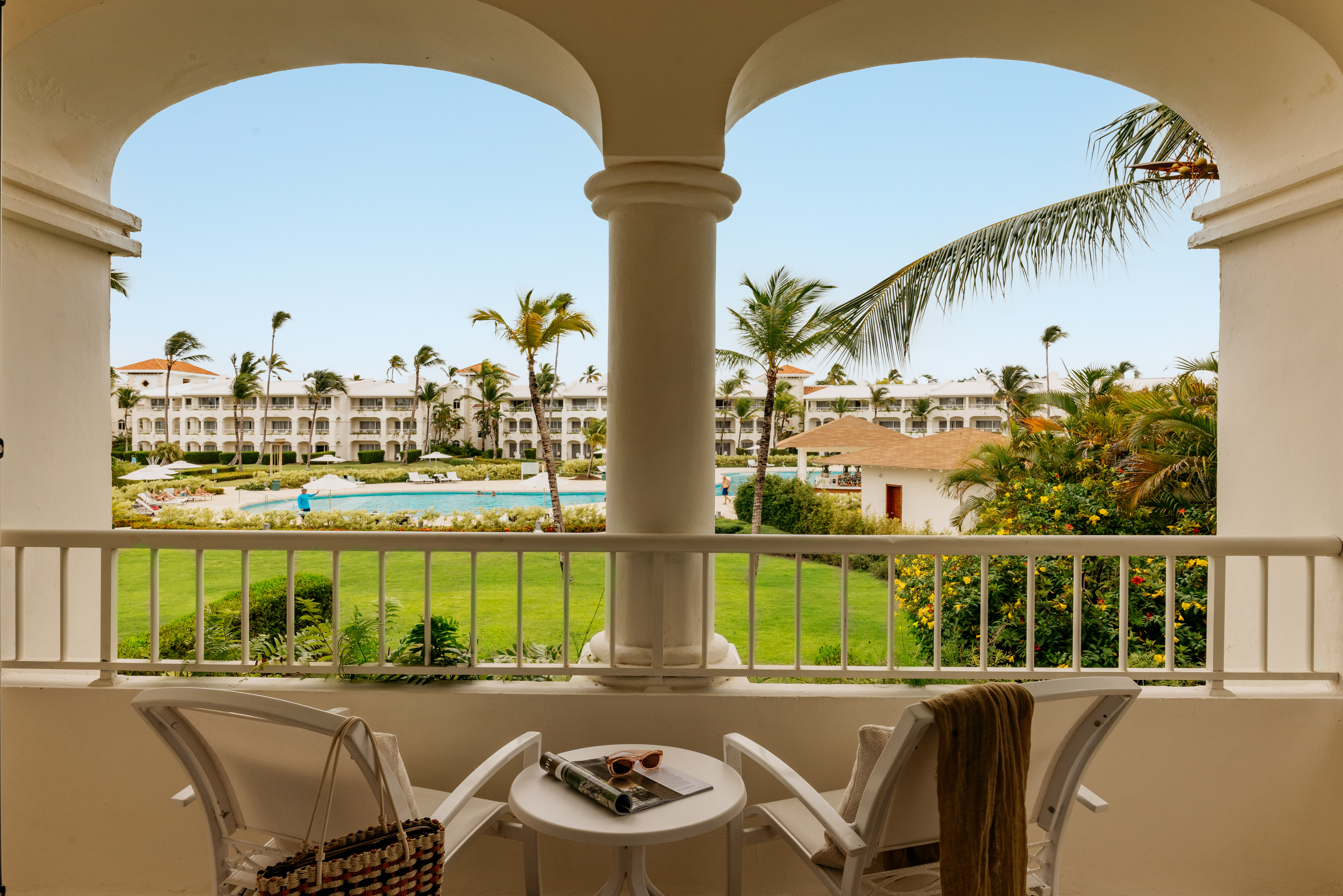 Tropical resort balcony overlooking pool, lush gardens, and white buildings.
