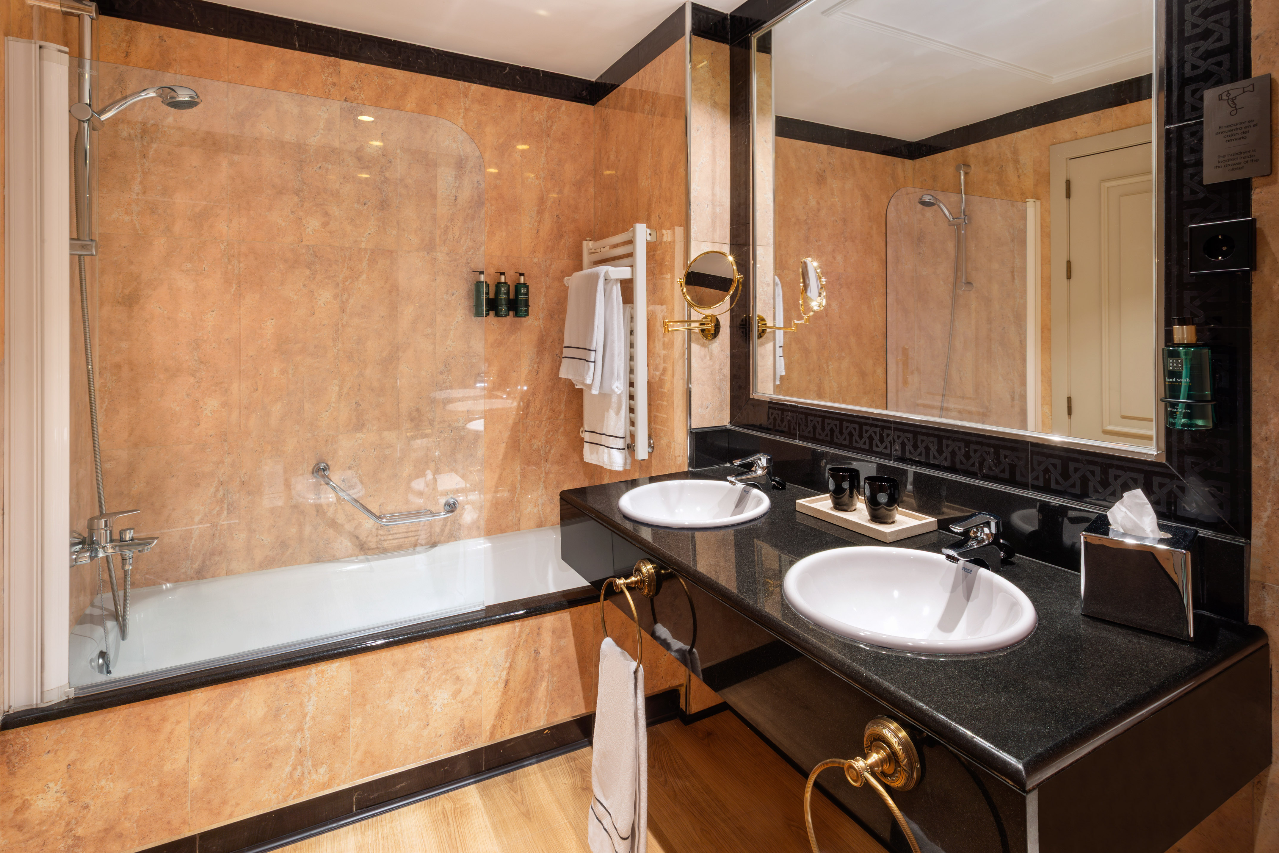 Modern hotel bathroom with double vanity, black granite counter, bathtub, and tiled walls.
