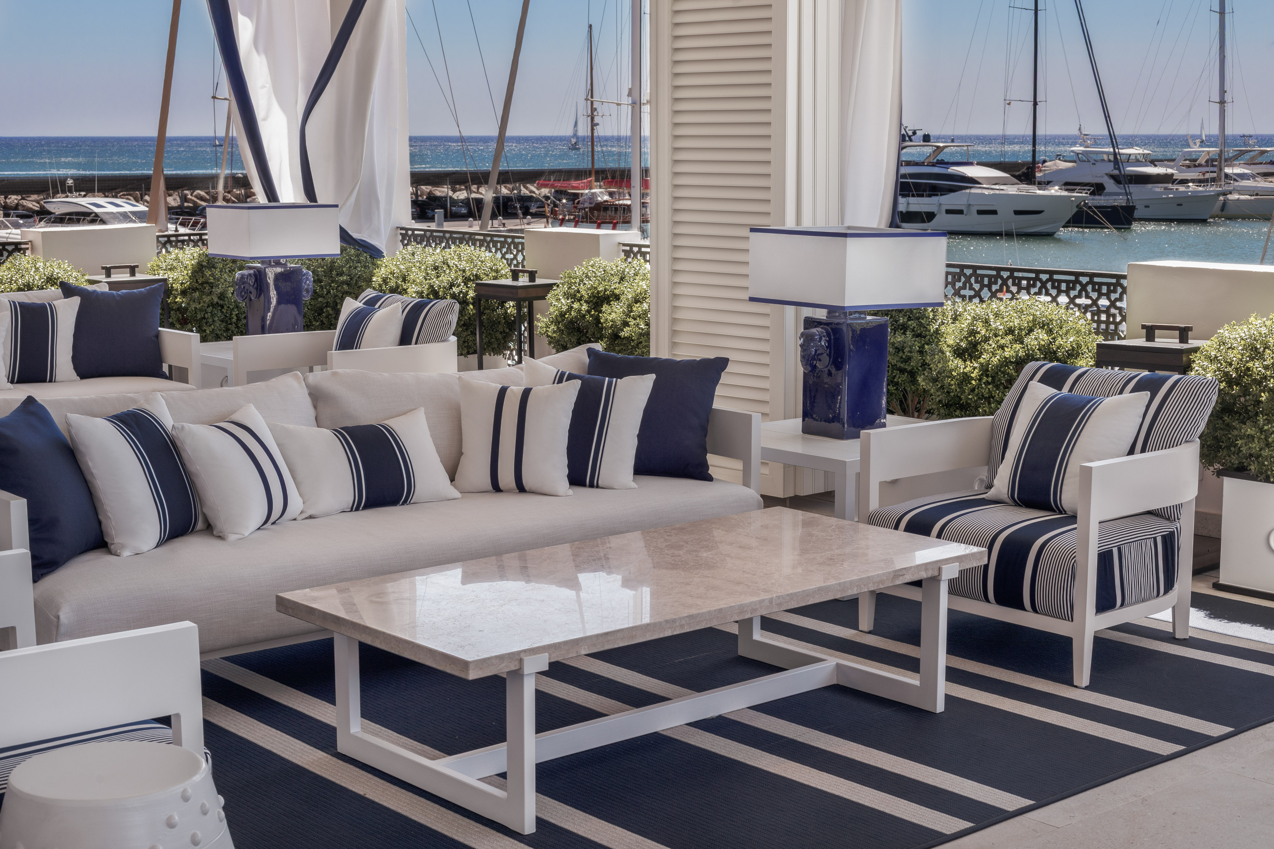 Coastal outdoor lounge with blue/white furniture, marble table, marina view.