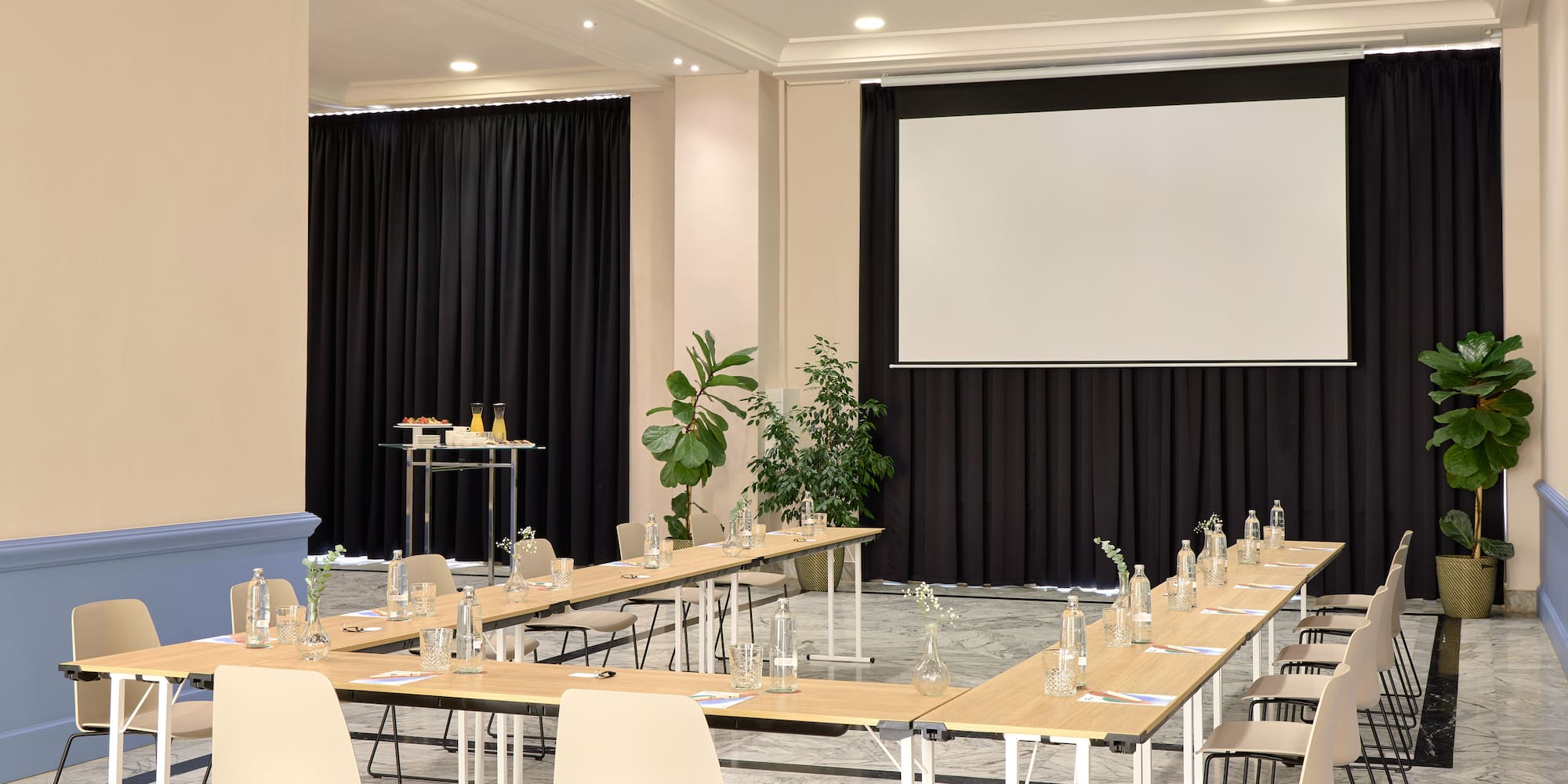 Sala de reuniones de hotel con mesas en U, proyector, pantalla y bebidas.
