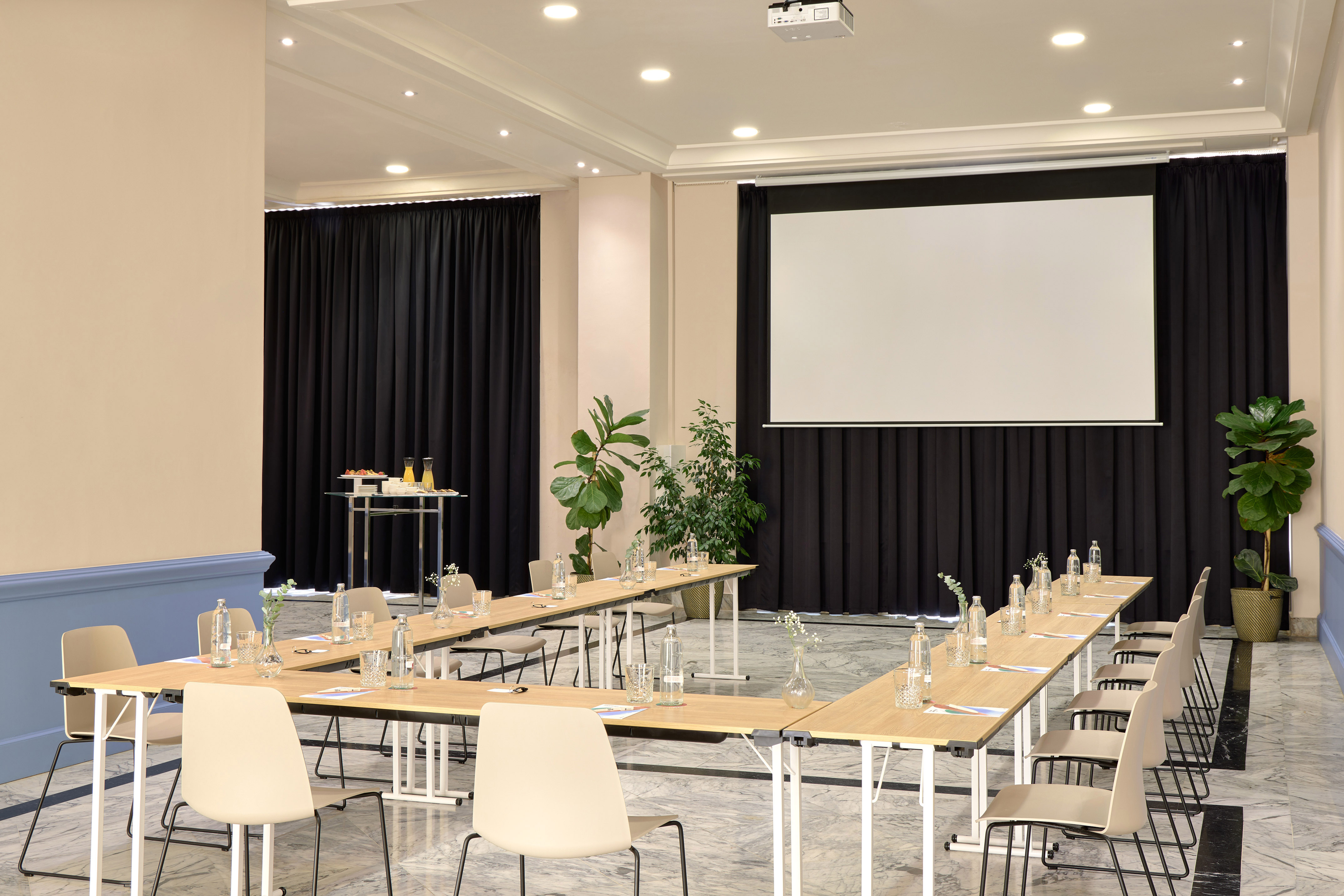 Sala de reuniones de hotel con mesas en U, proyector, pantalla y bebidas.