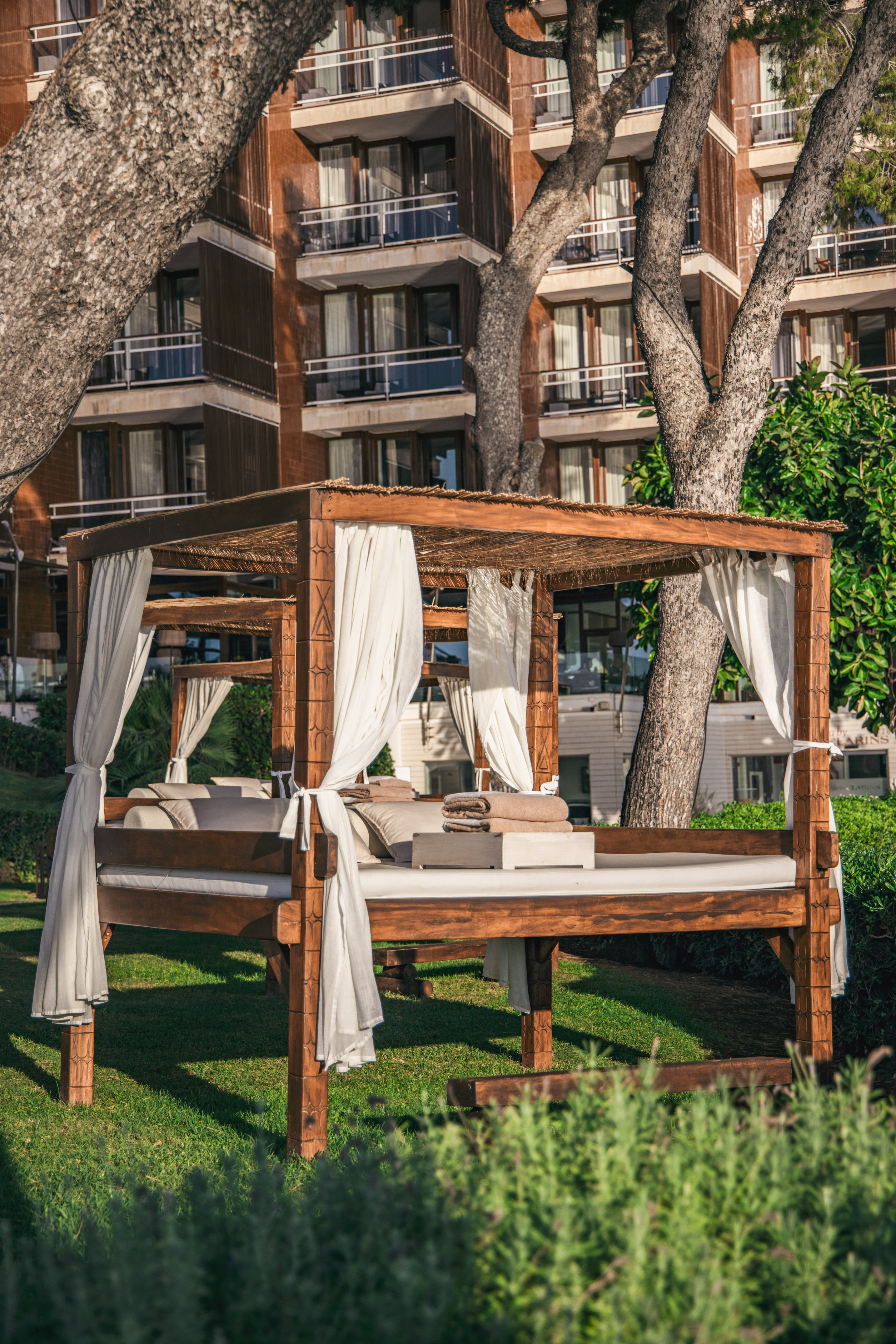 Luxury resort daybed with white drapes on green grass, hotel building and trees.