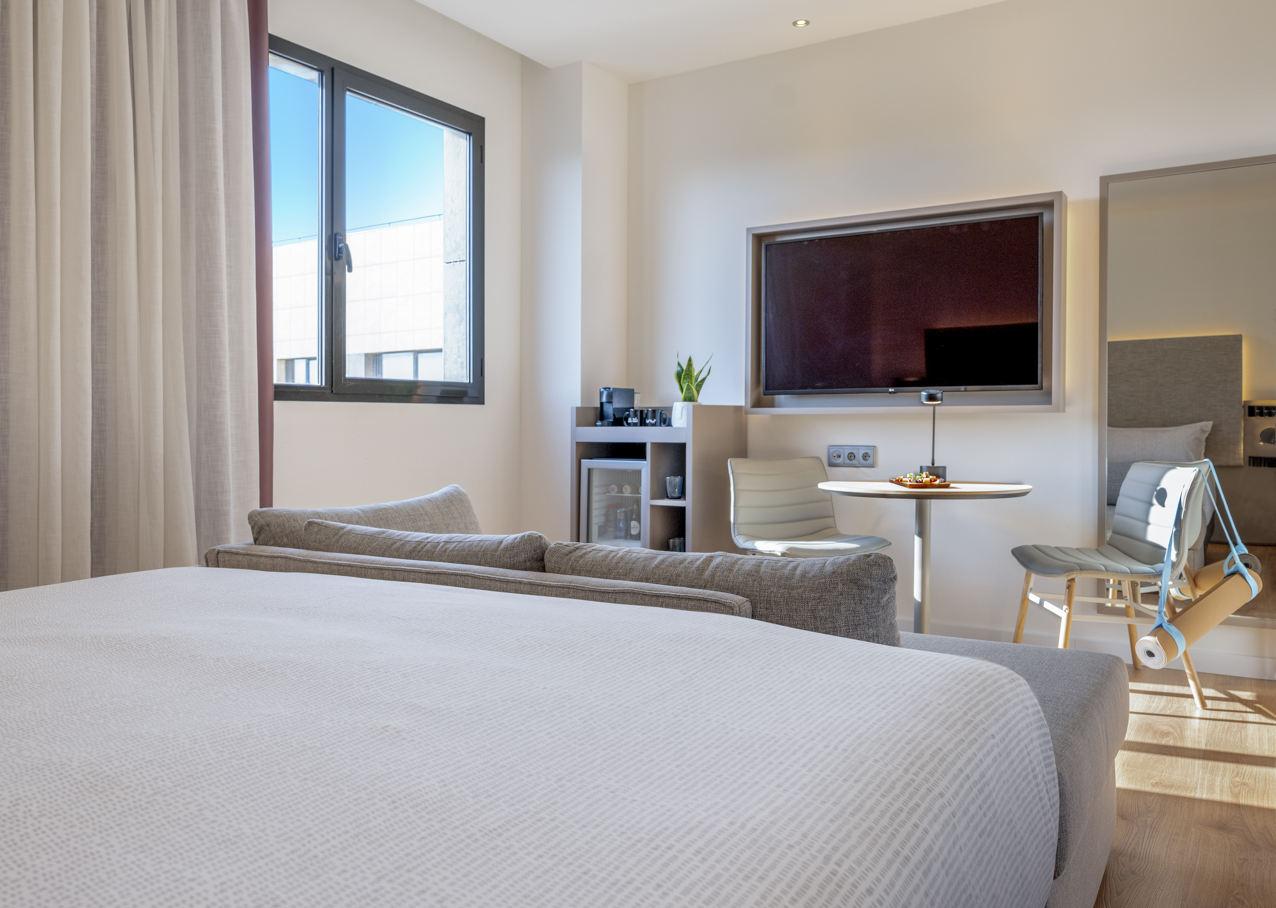 Bright, modern hotel room with bed, sofa, TV, minibar, and yoga mat.