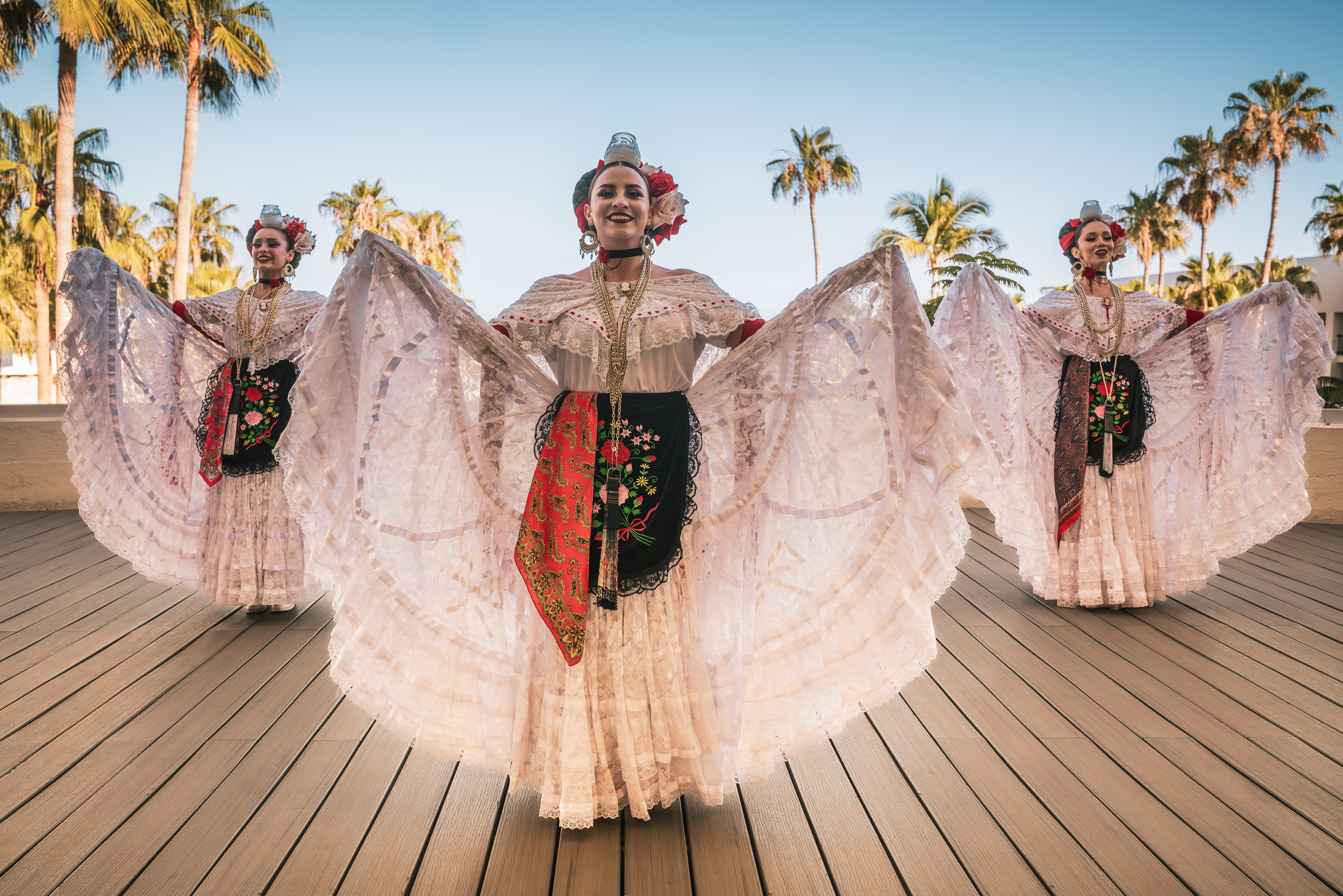 Three Mexican folkloric dancers in traditional lace dresses, palm trees.