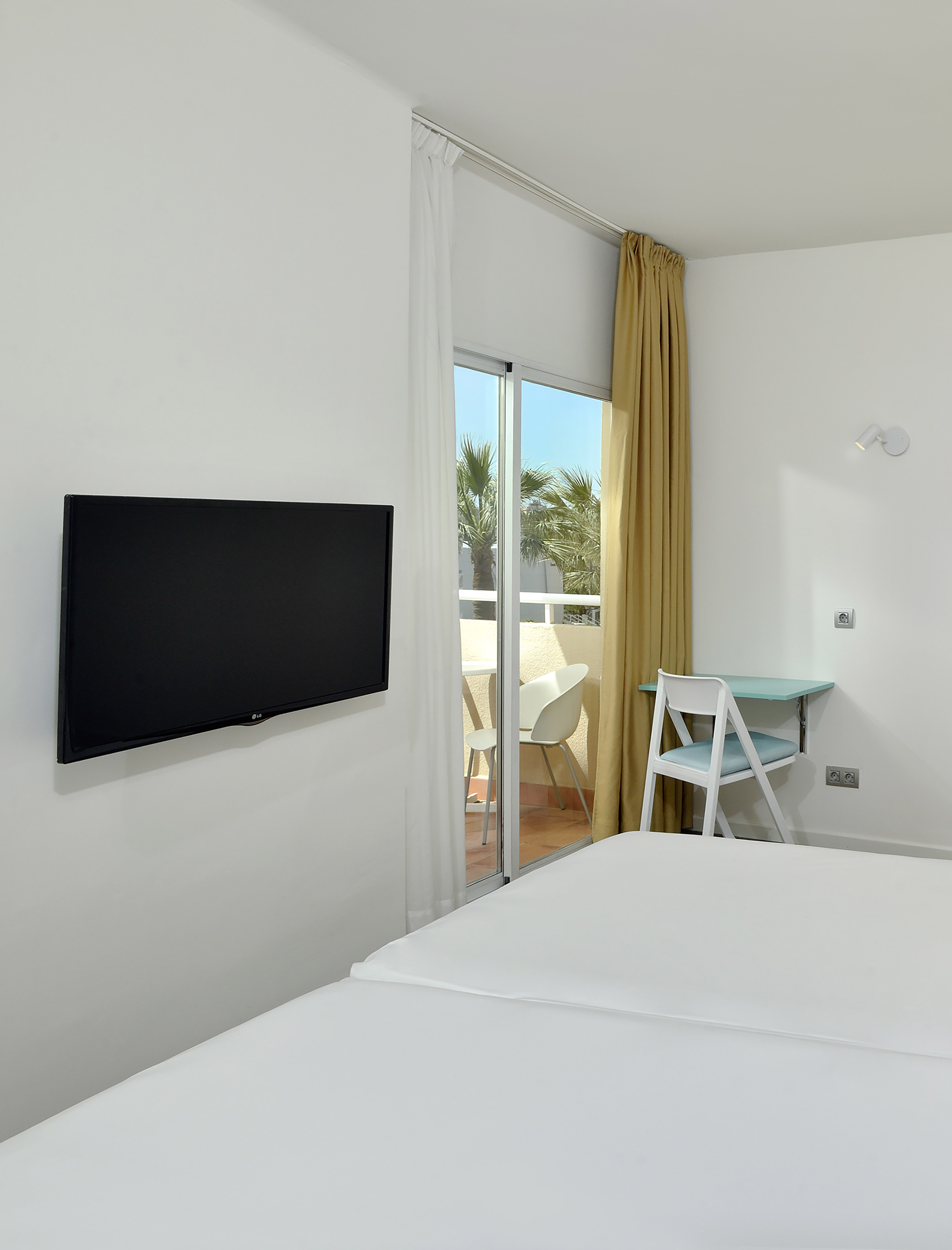 Hotel bedroom with TV, white bed, light blue desk, and balcony with palm trees.