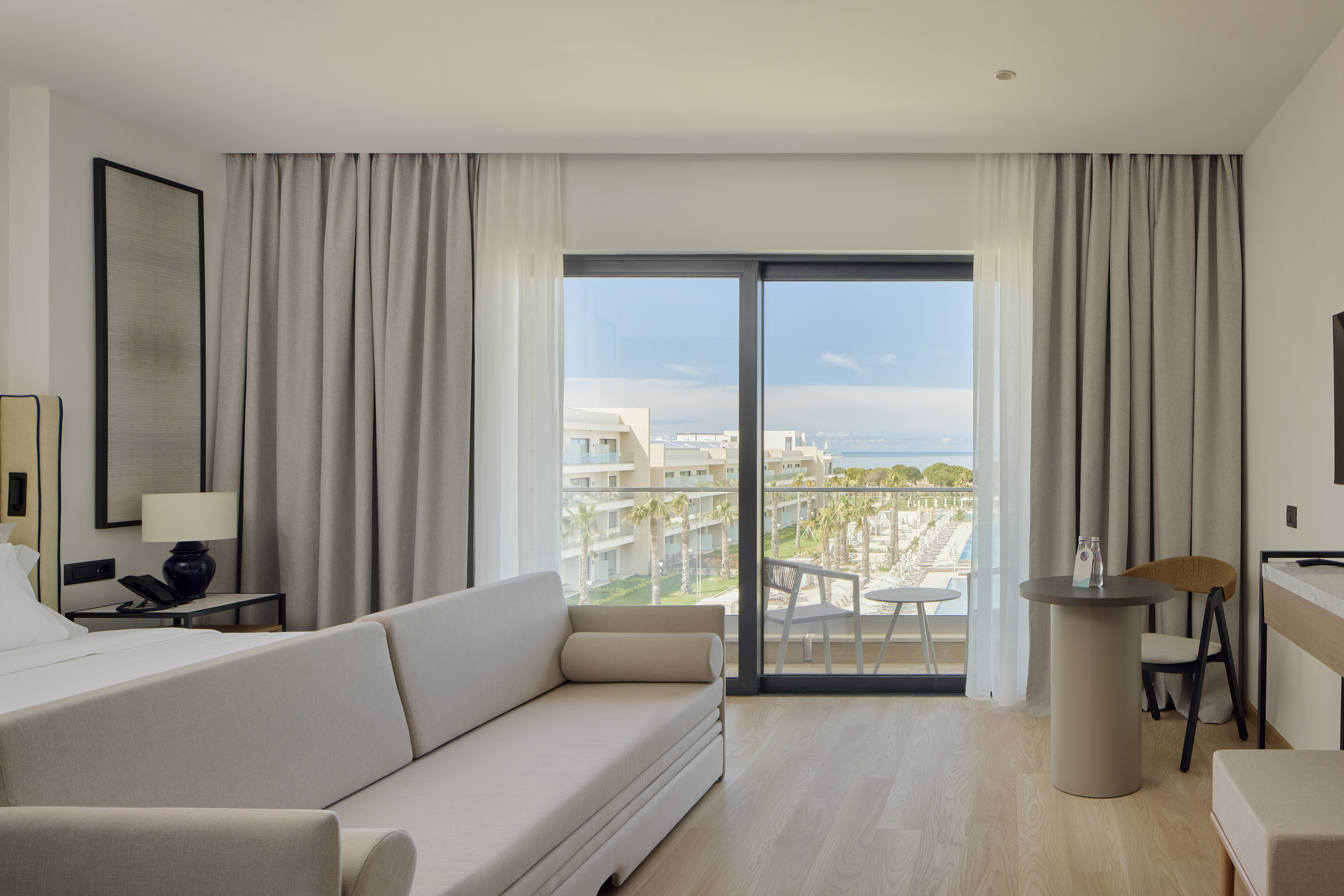 Modern resort suite with ocean view balcony, sofa, pool, and palm trees.