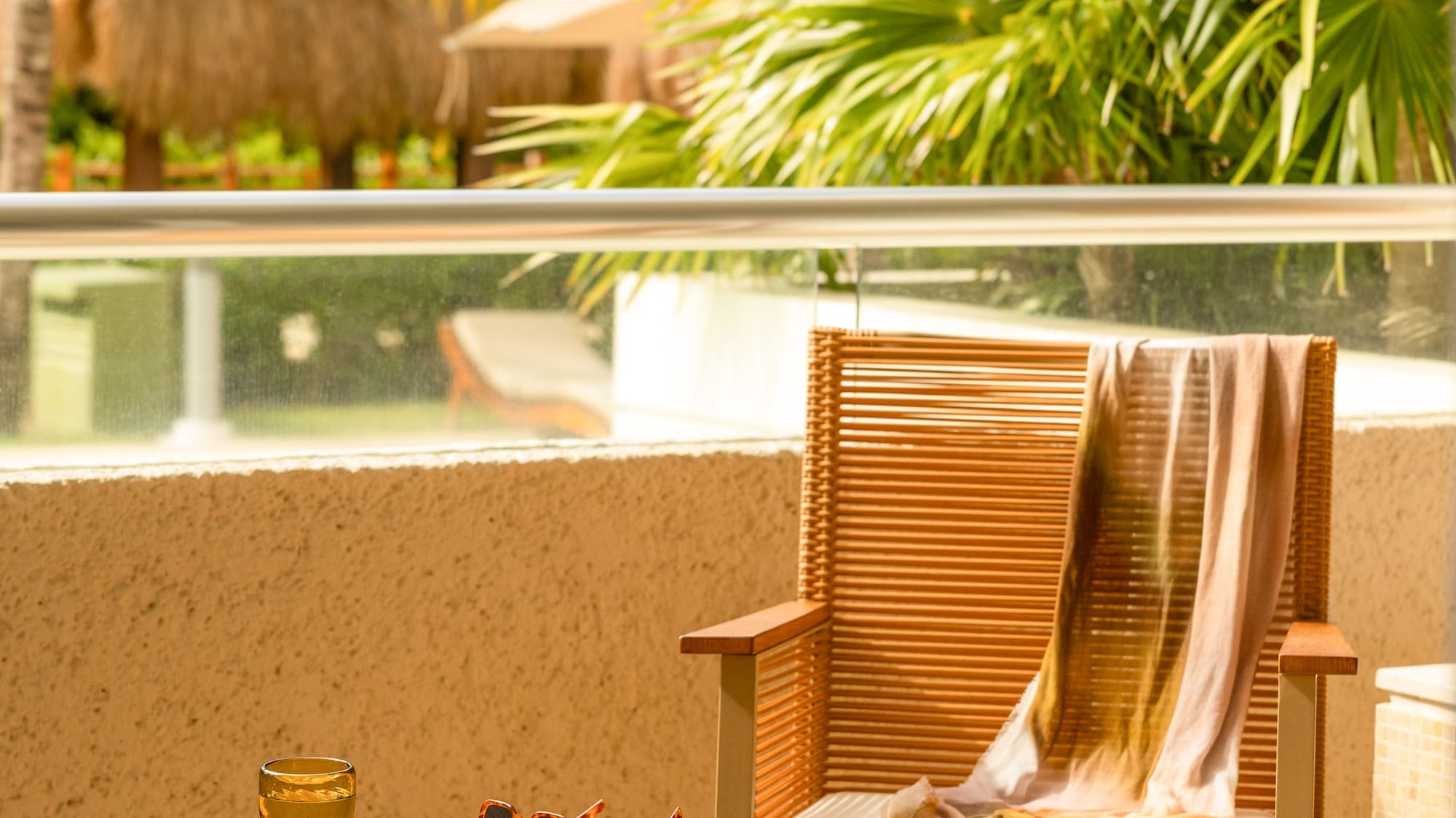 Tropical resort balcony with wicker chair, table, drink, sunglasses, palm trees.
