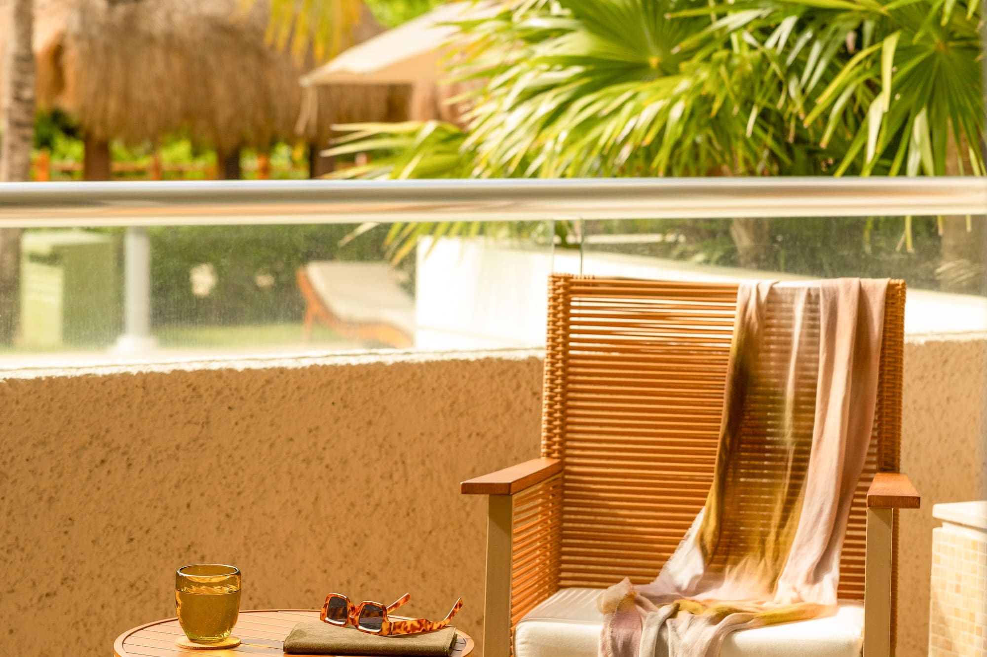 Tropical resort balcony with wicker chair, table, drink, sunglasses, palm trees.