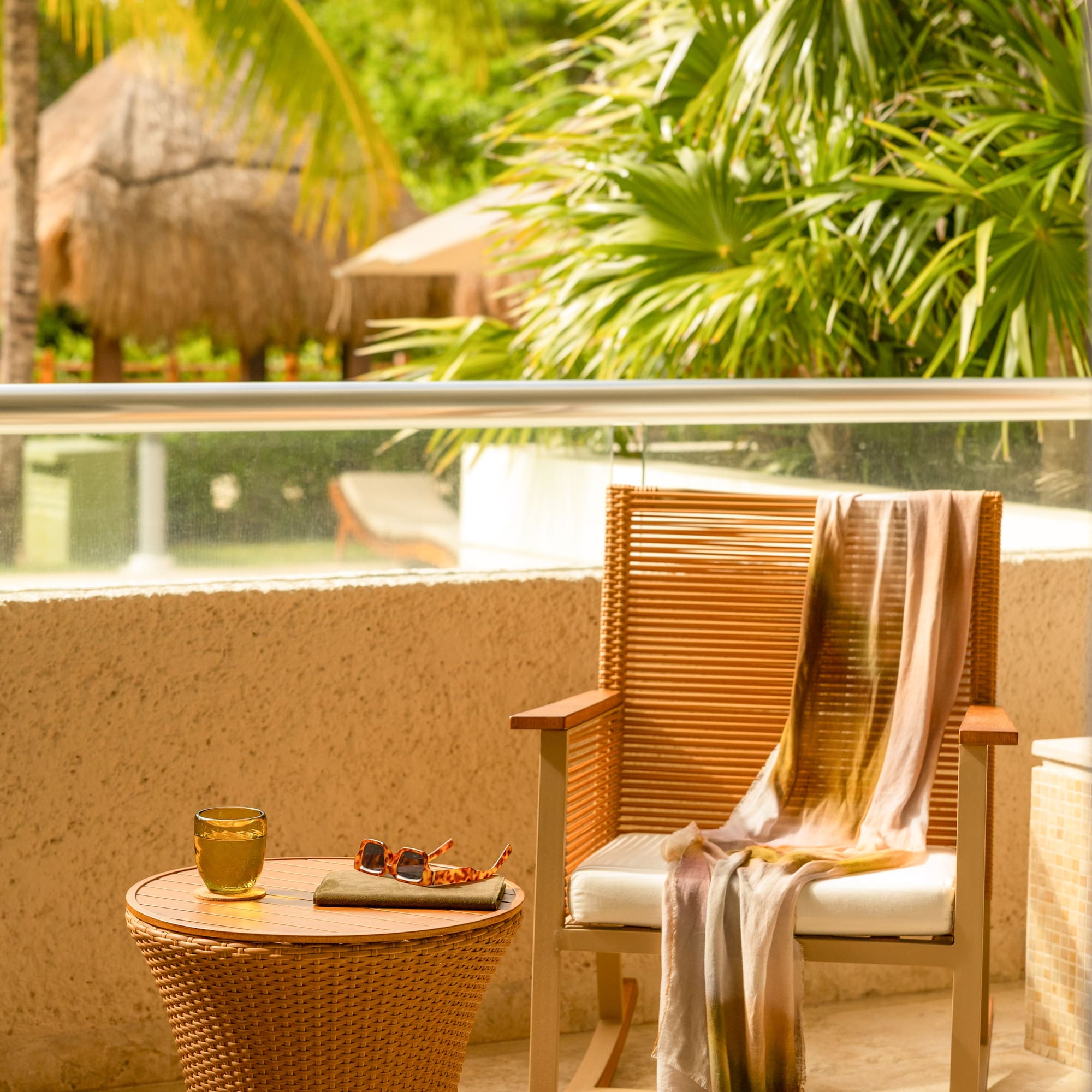 Tropical resort balcony with wicker chair, table, drink, sunglasses, palm trees.