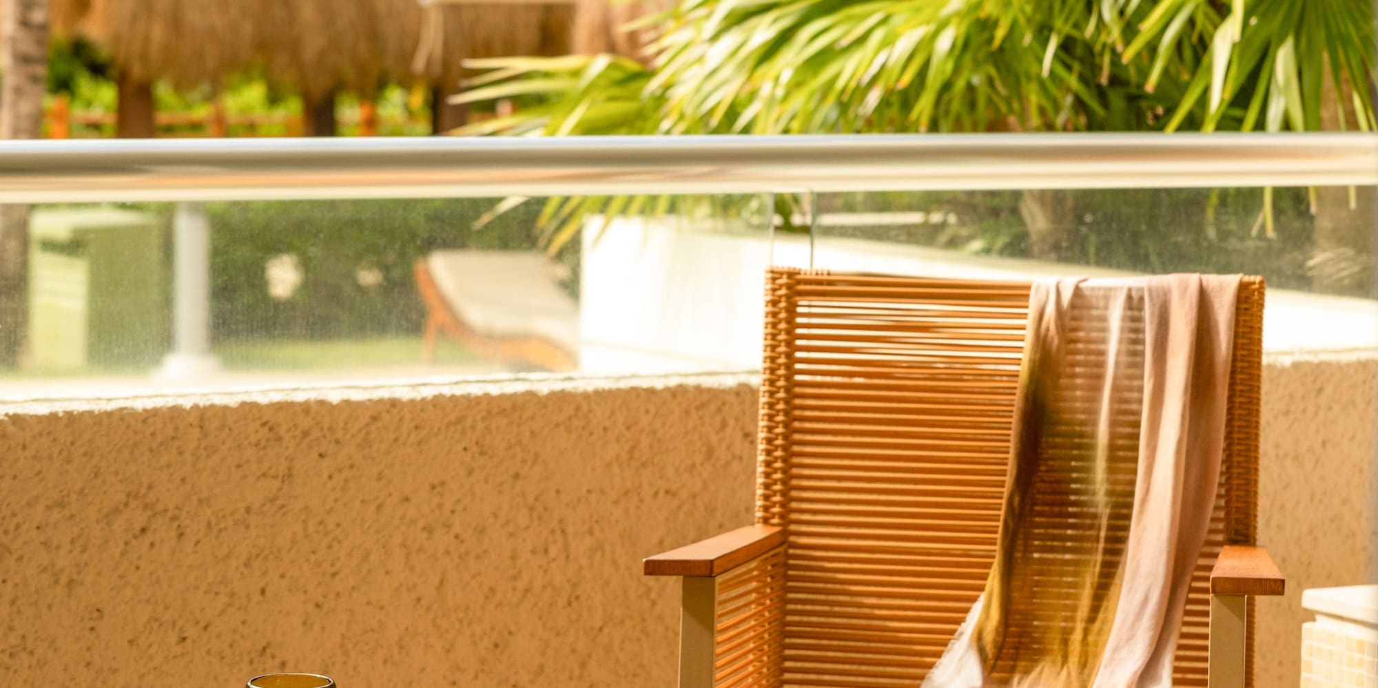Tropical resort balcony with wicker chair, table, drink, sunglasses, palm trees.
