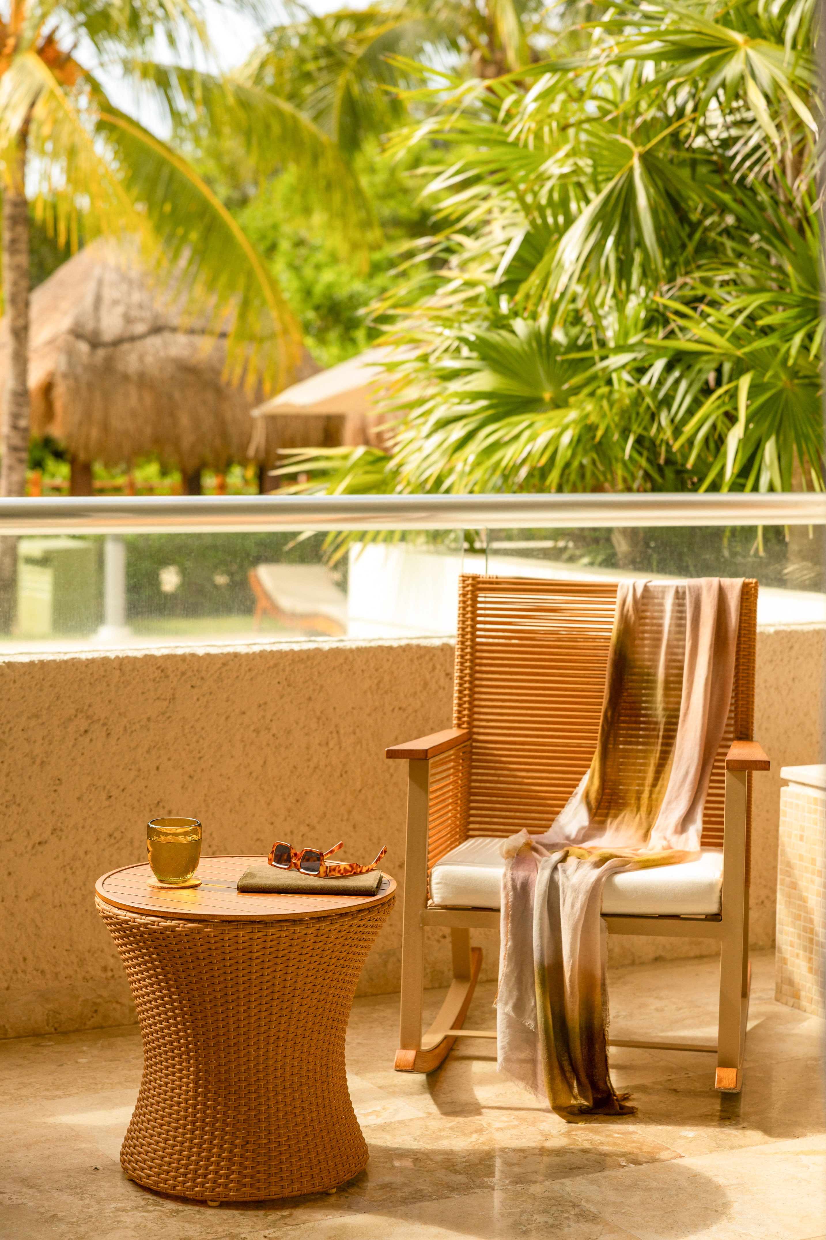 Tropical resort balcony with wicker chair, table, drink, sunglasses, palm trees.
