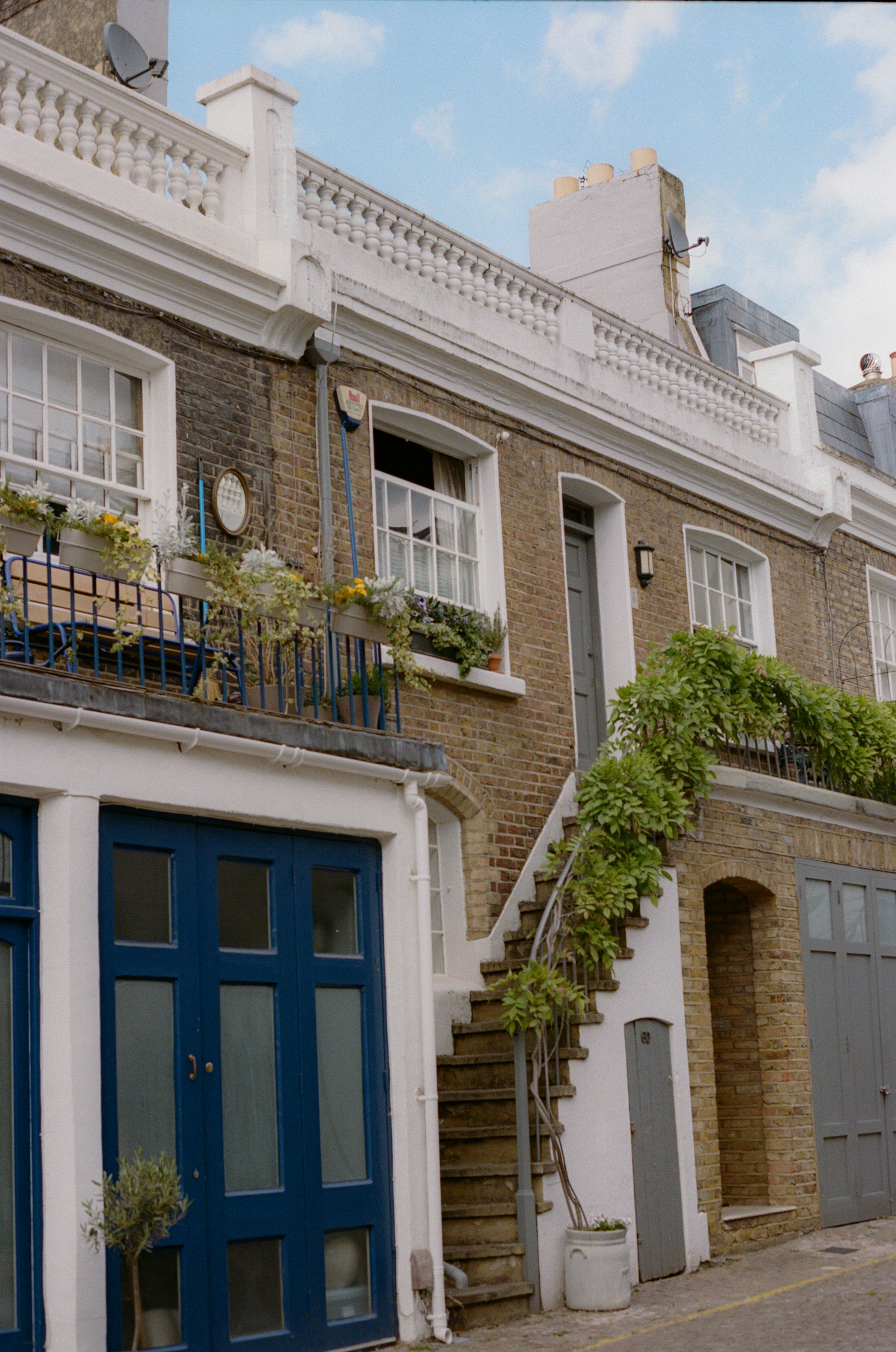 Fachada de ladrillo, puertas azules, balcones con flores y escaleras con vegetación.