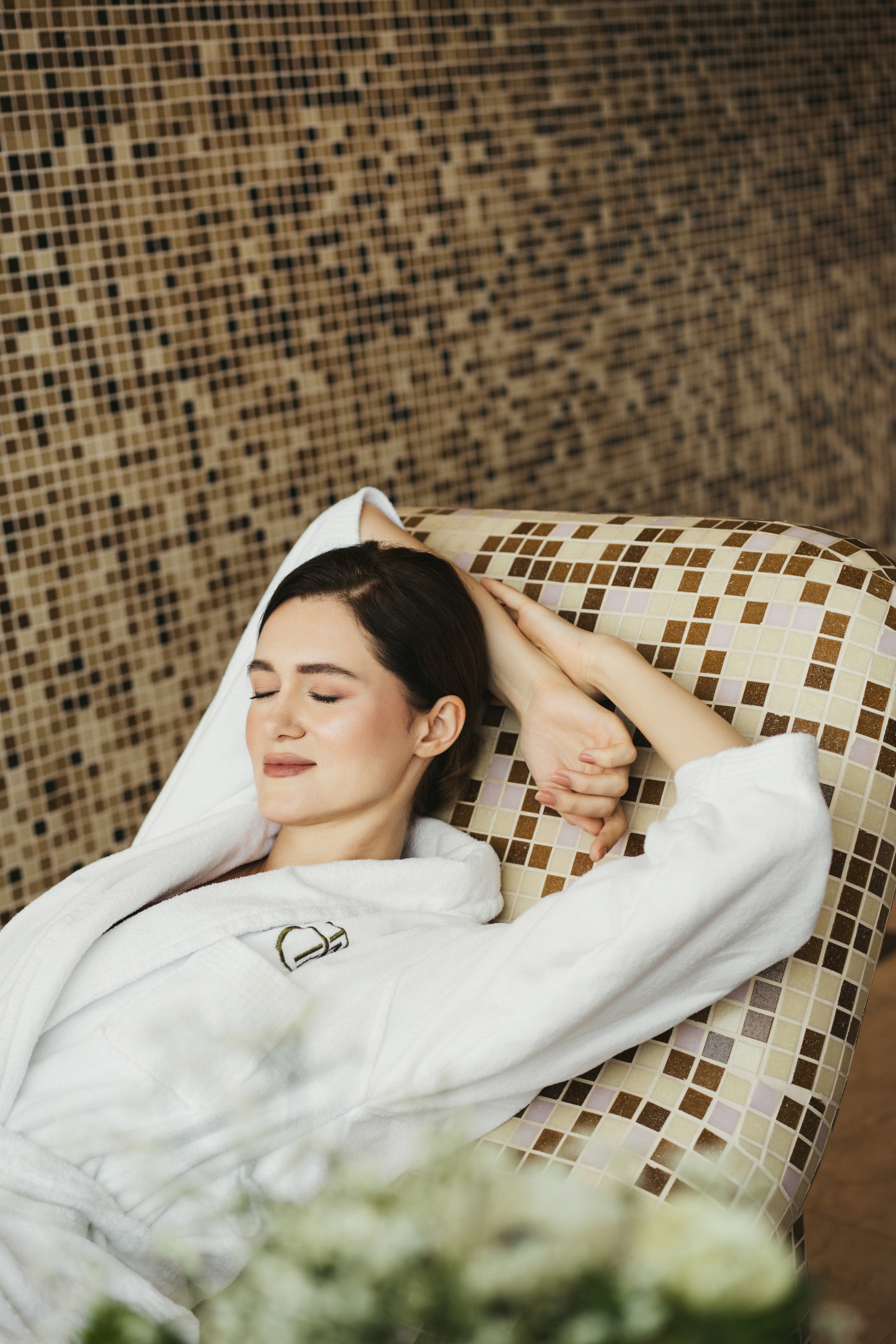 Serene woman in white bathrobe relaxing on mosaic spa lounger, eyes closed.