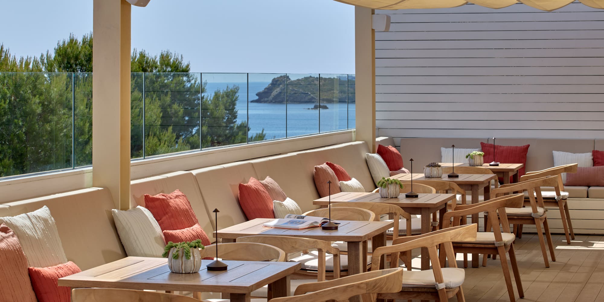 Sunny outdoor dining terrace with sea view, wooden tables, and cushioned seating.