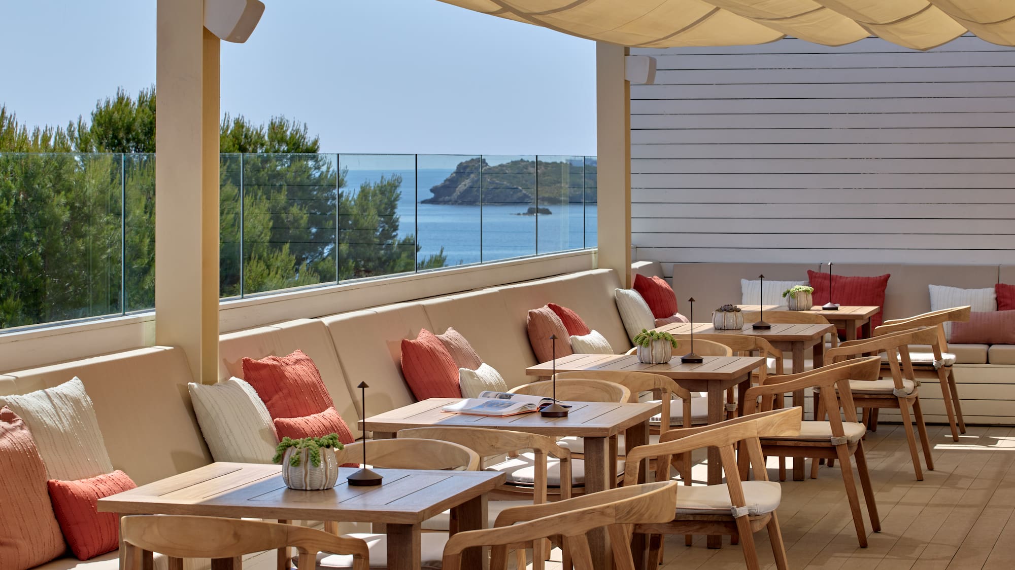 Sunny outdoor dining terrace with sea view, wooden tables, and cushioned seating.