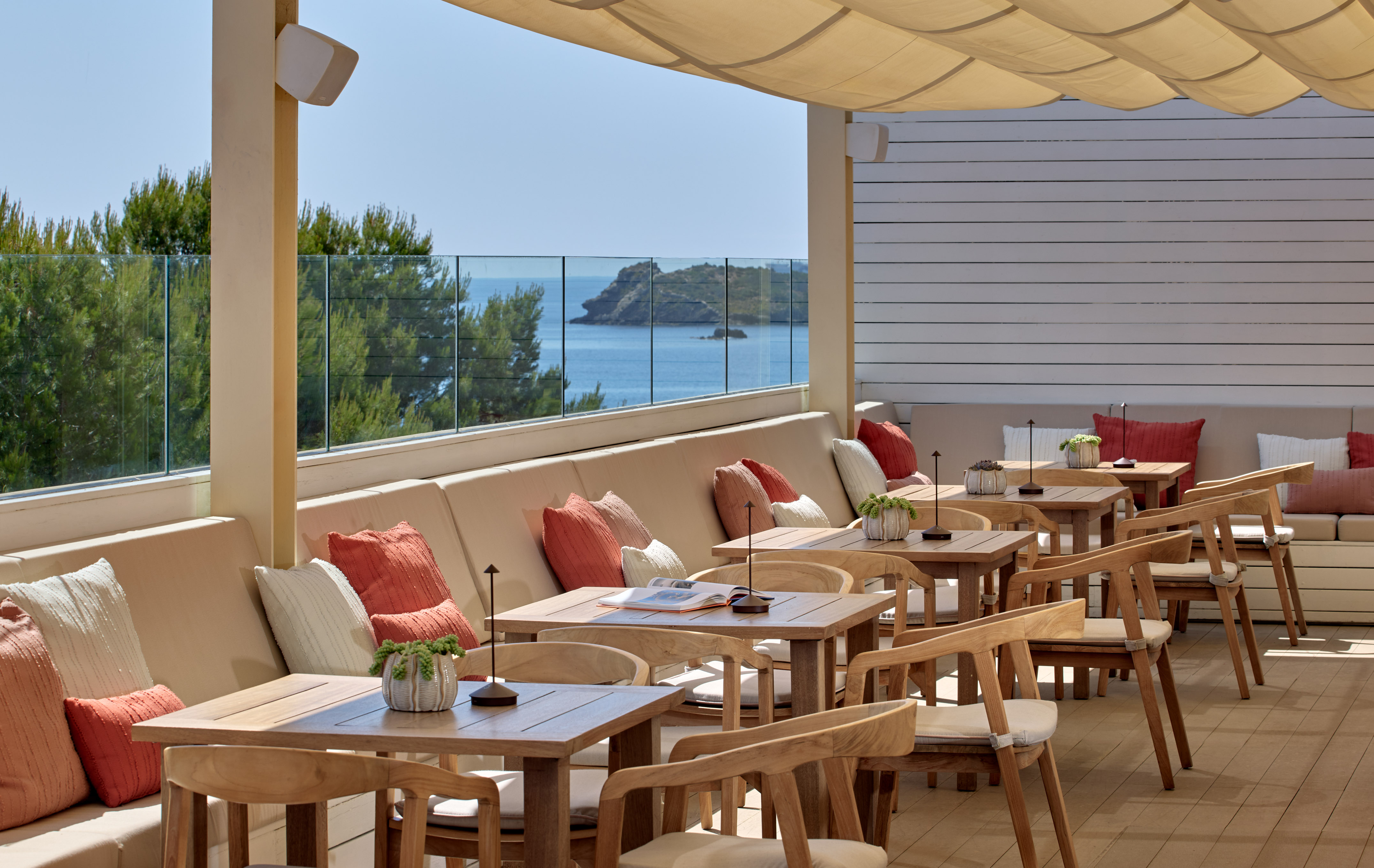Sunny outdoor dining terrace with sea view, wooden tables, and cushioned seating.