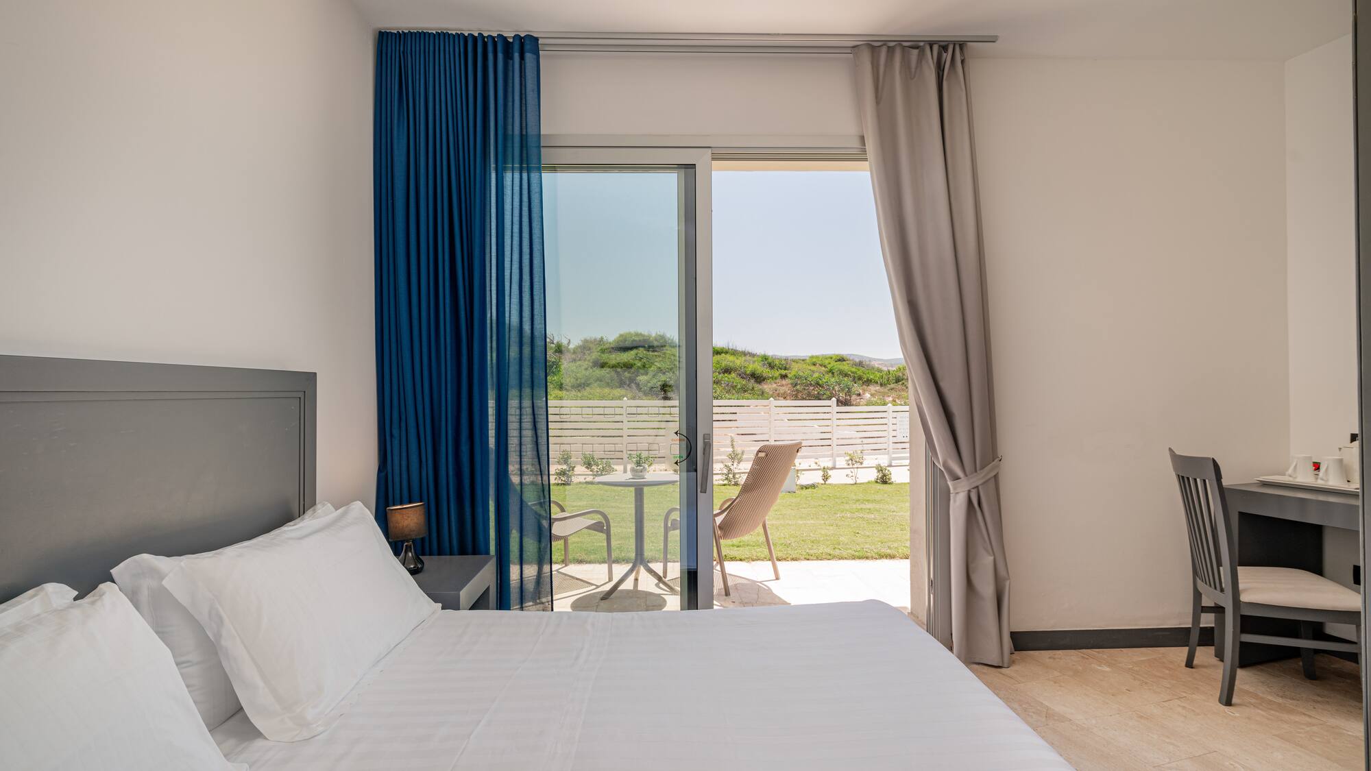 Bright hotel bedroom with white bed, patio access, and garden view.