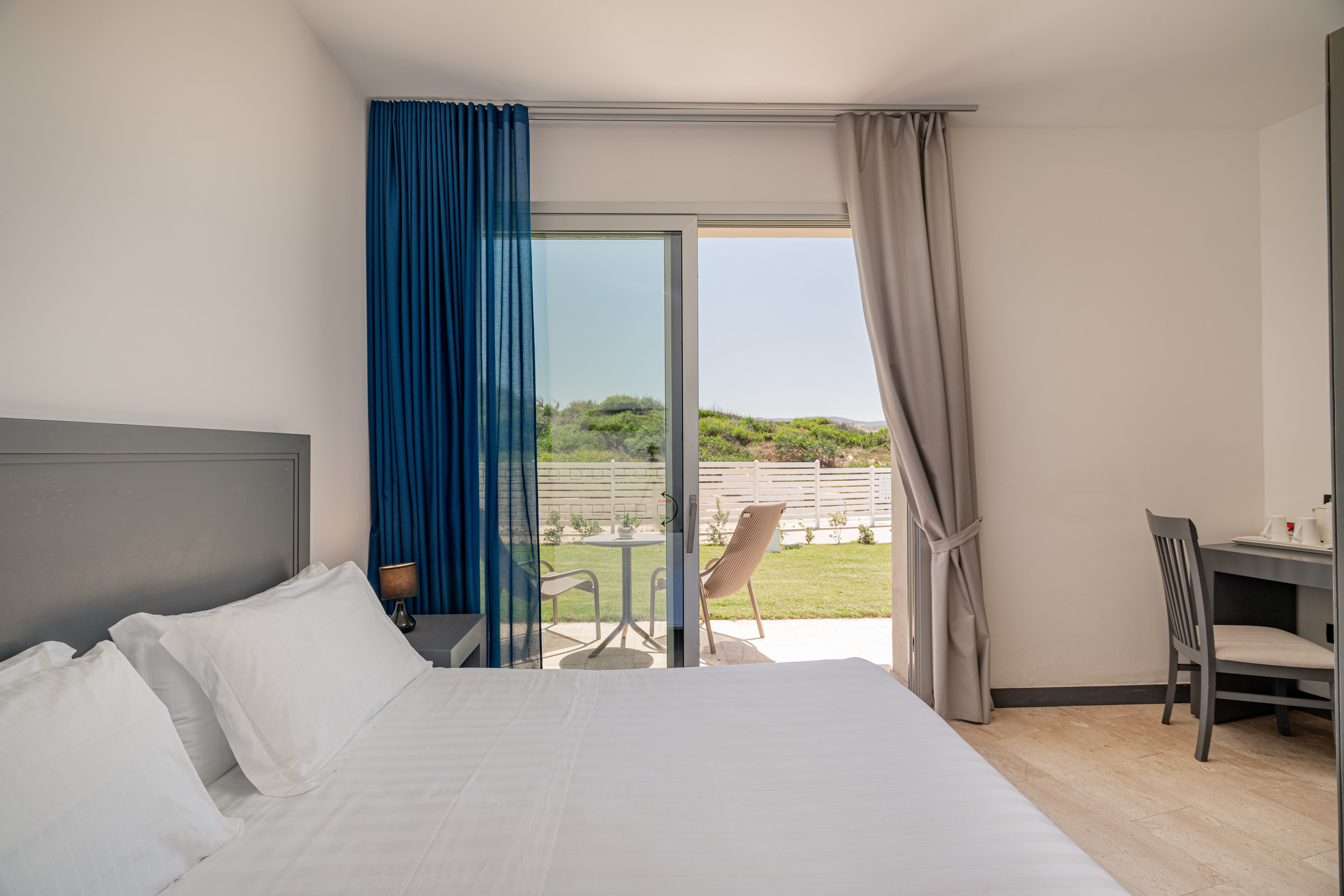 Bright hotel bedroom with white bed, patio access, and garden view.