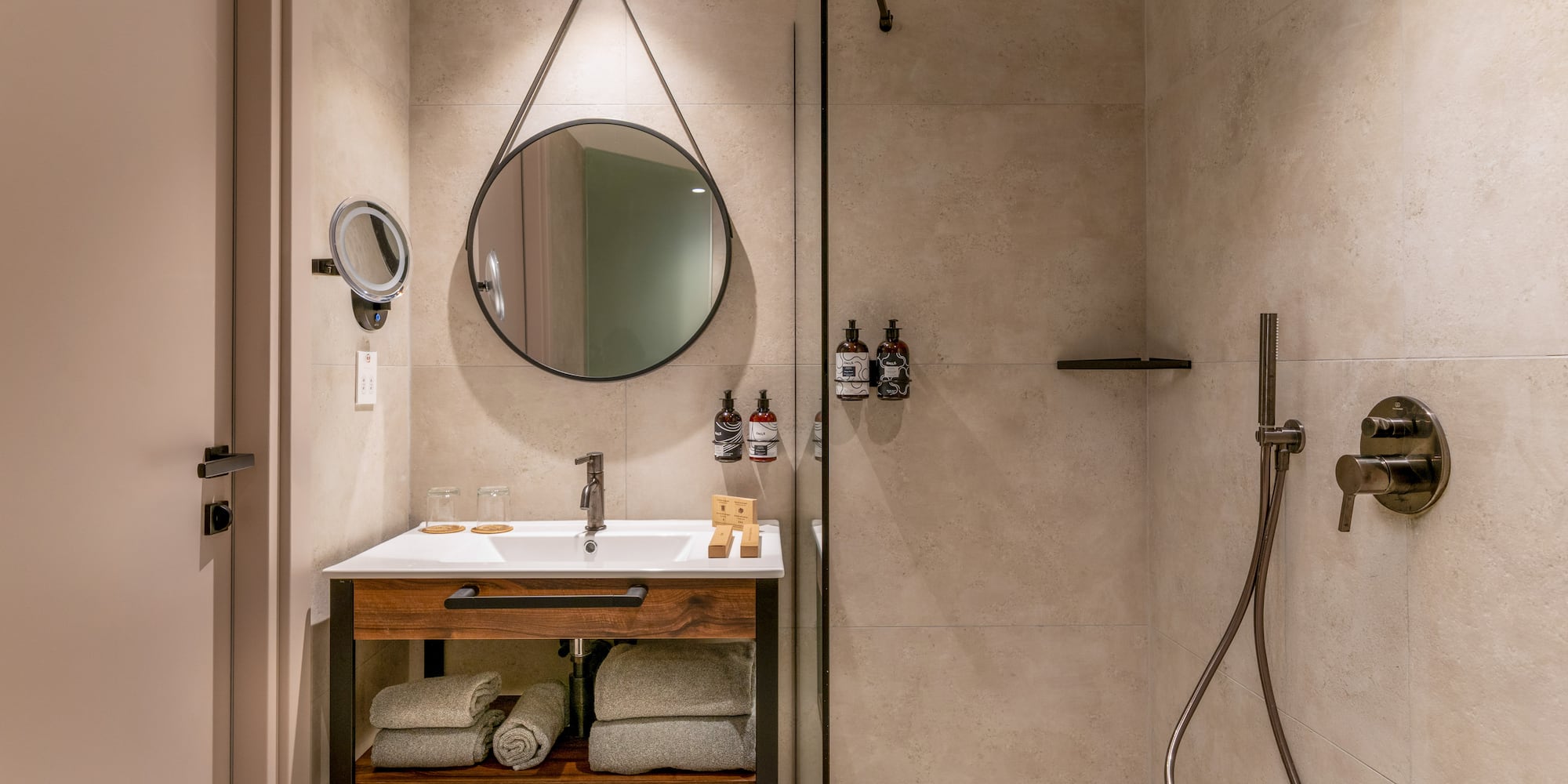 Modern hotel bathroom with wood vanity, round mirror, and walk-in shower.