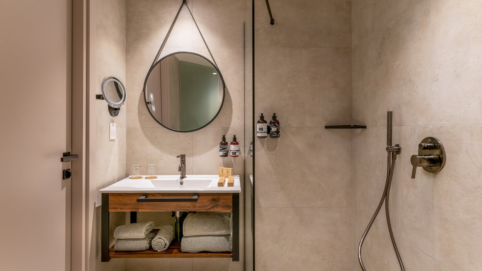 Modern hotel bathroom with wood vanity, round mirror, and walk-in shower.