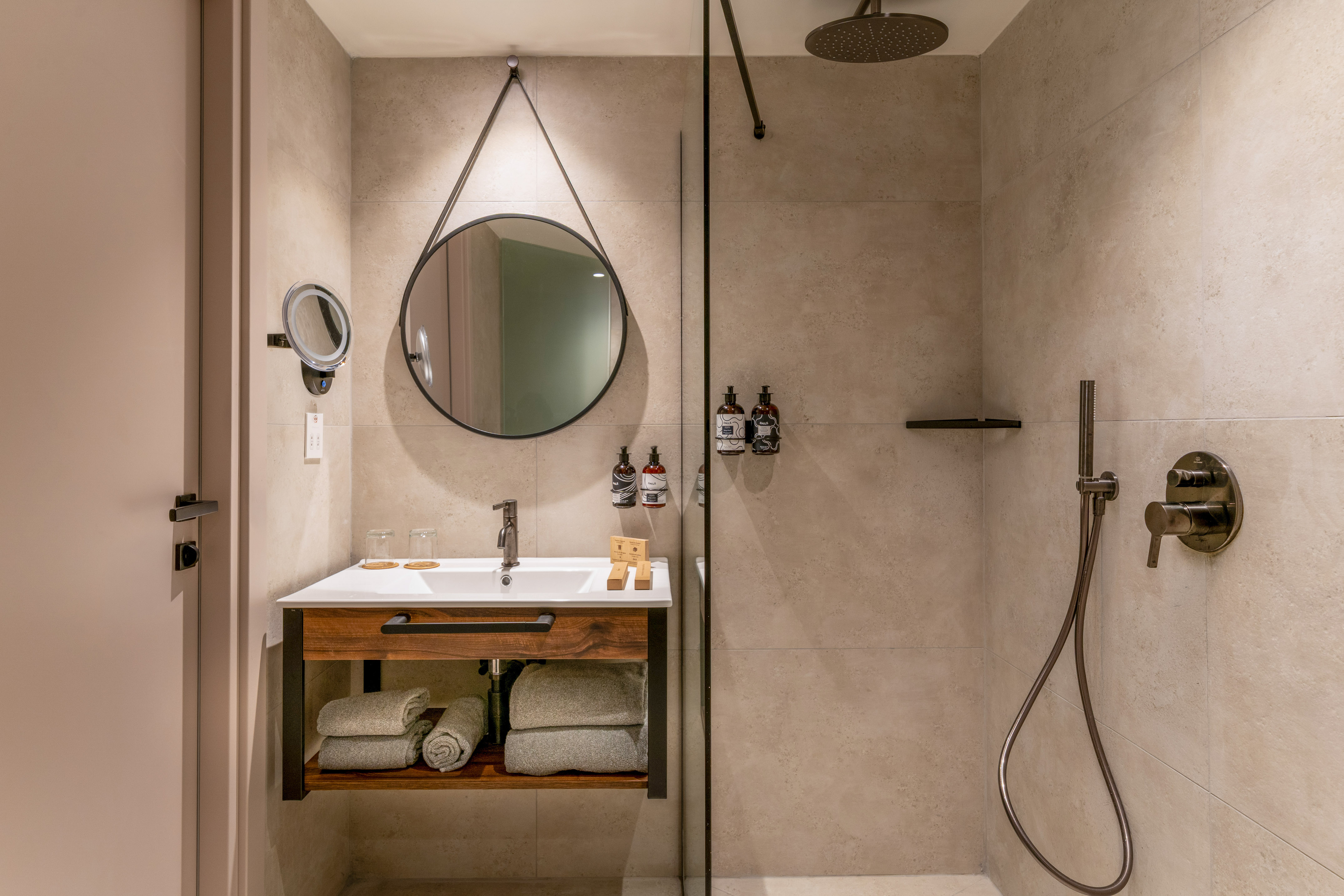 Modern hotel bathroom with wood vanity, round mirror, and walk-in shower.