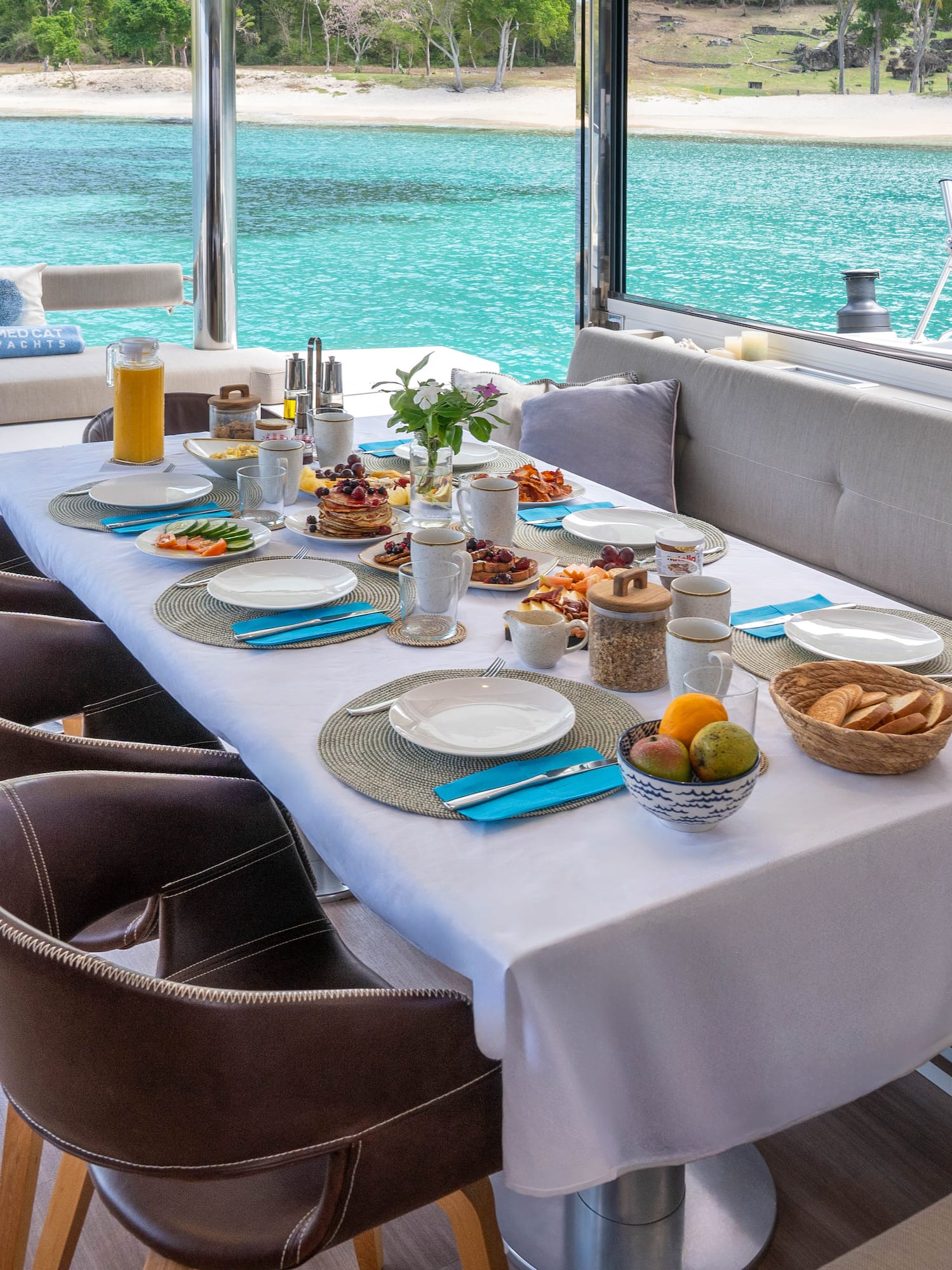 Breakfast table on a catamaran deck, full spread, tropical ocean and beach.