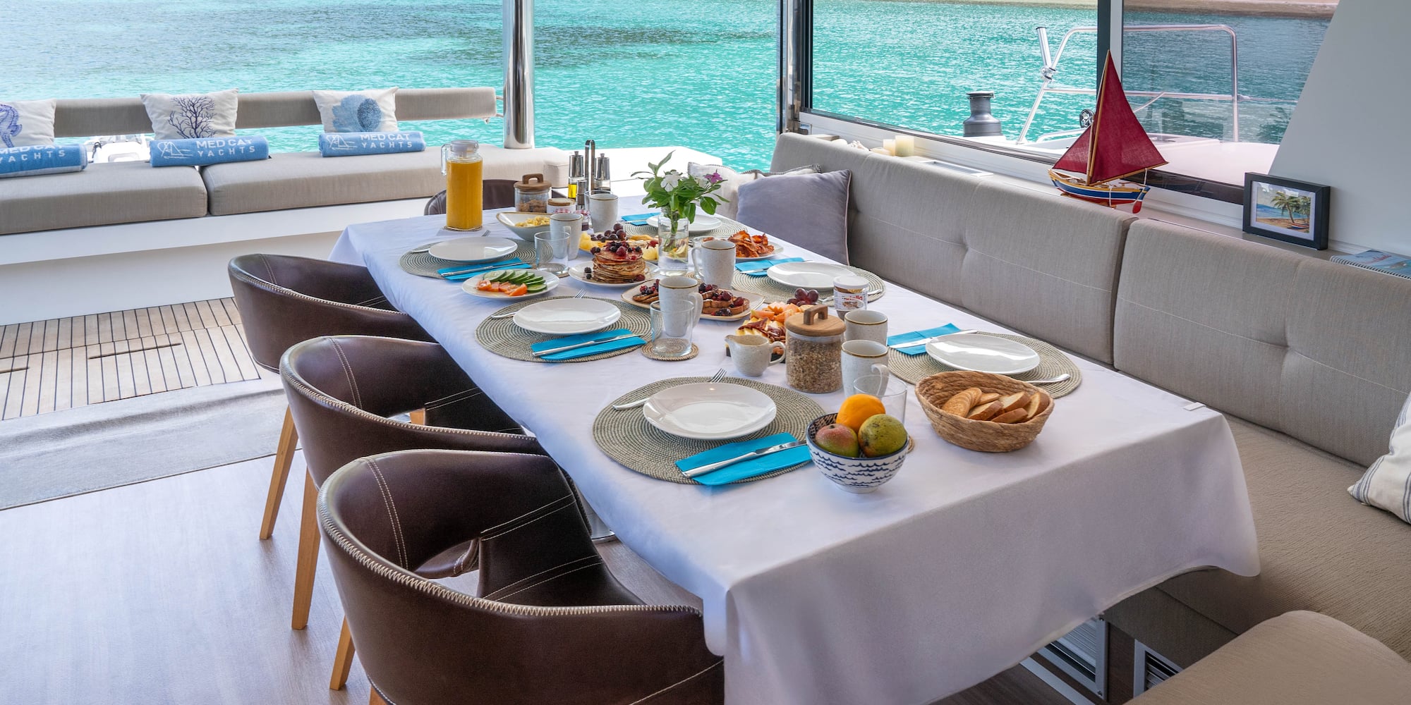 Breakfast table on a catamaran deck, full spread, tropical ocean and beach.