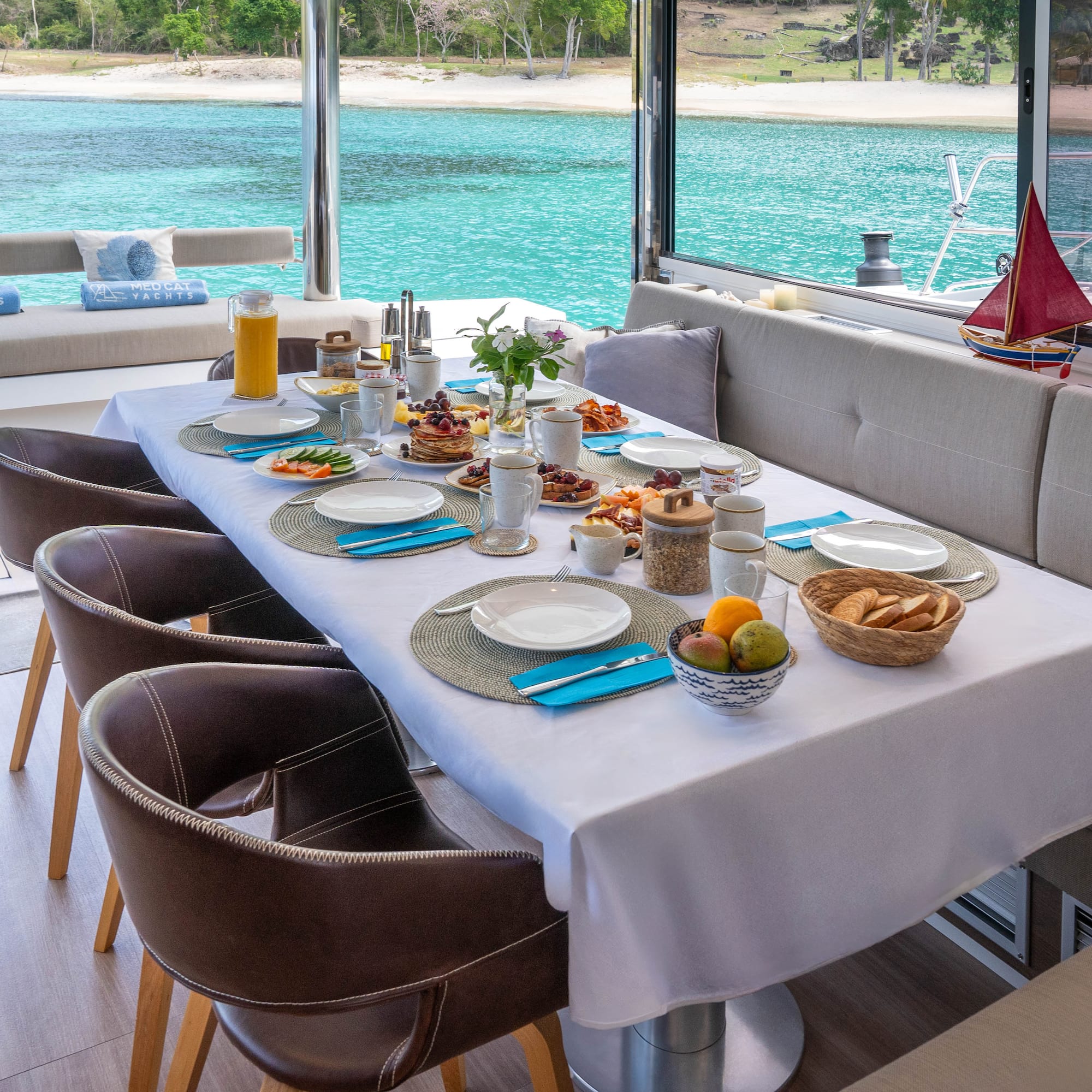 Breakfast table on a catamaran deck, full spread, tropical ocean and beach.