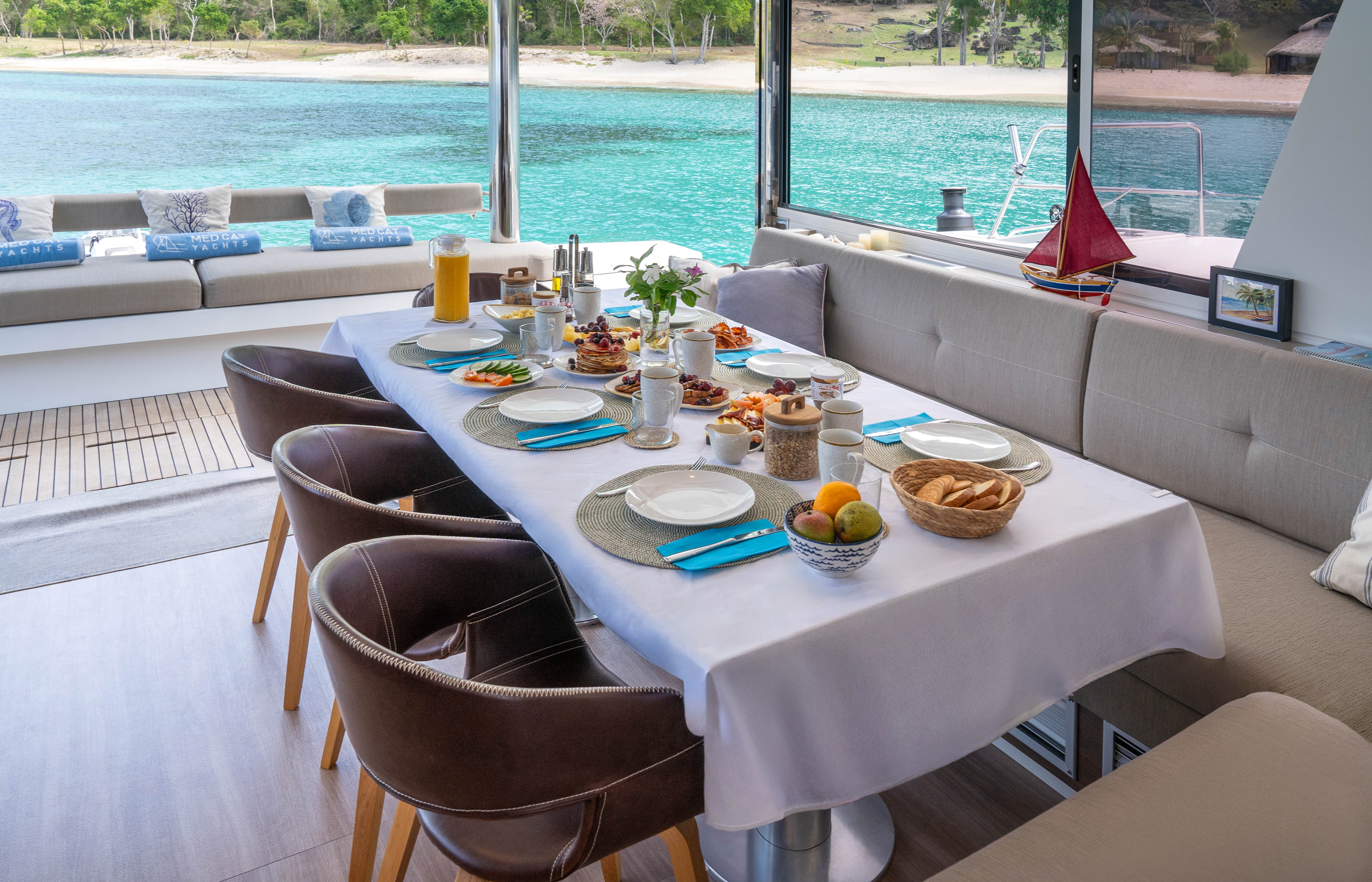 Breakfast table on a catamaran deck, full spread, tropical ocean and beach.