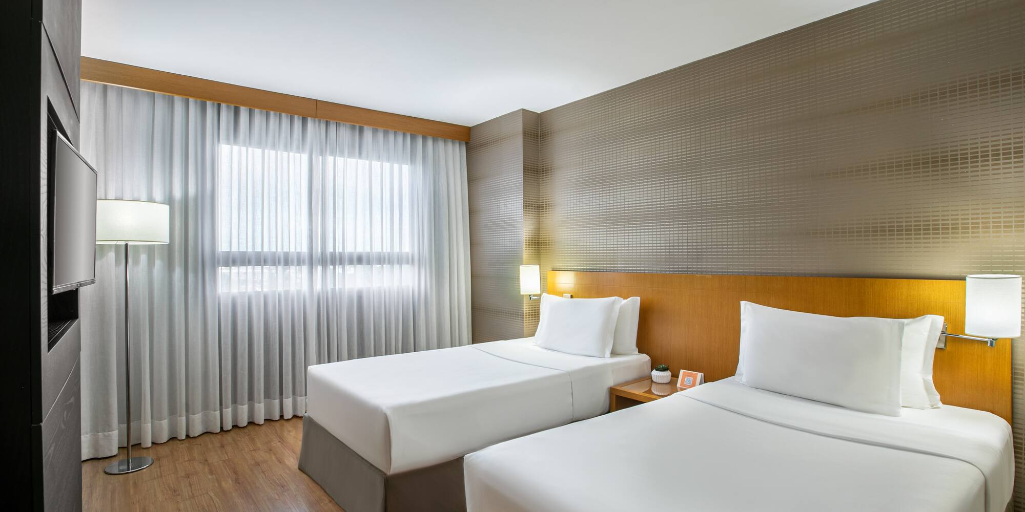 Modern hotel guest room with two twin beds, wood decor, and bright window.