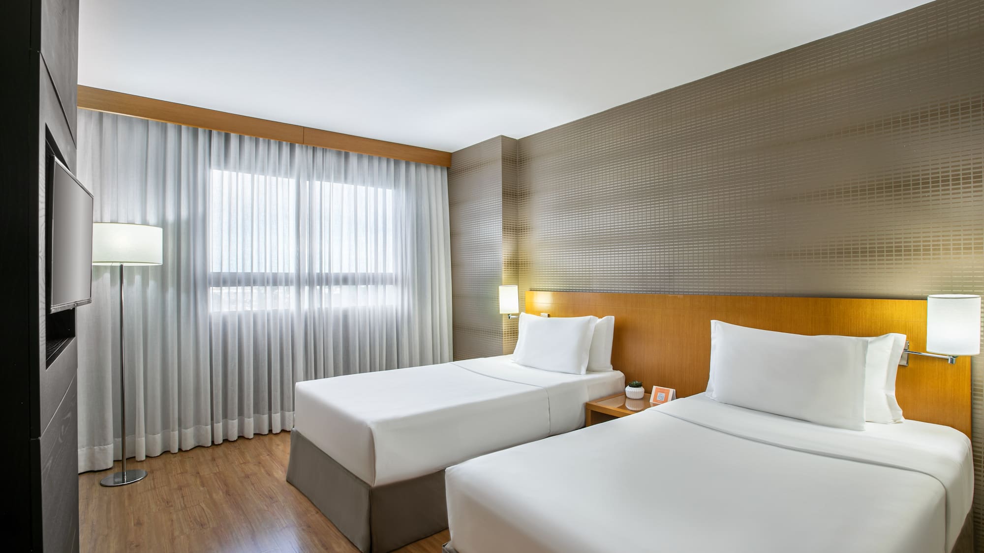 Modern hotel guest room with two twin beds, wood decor, and bright window.