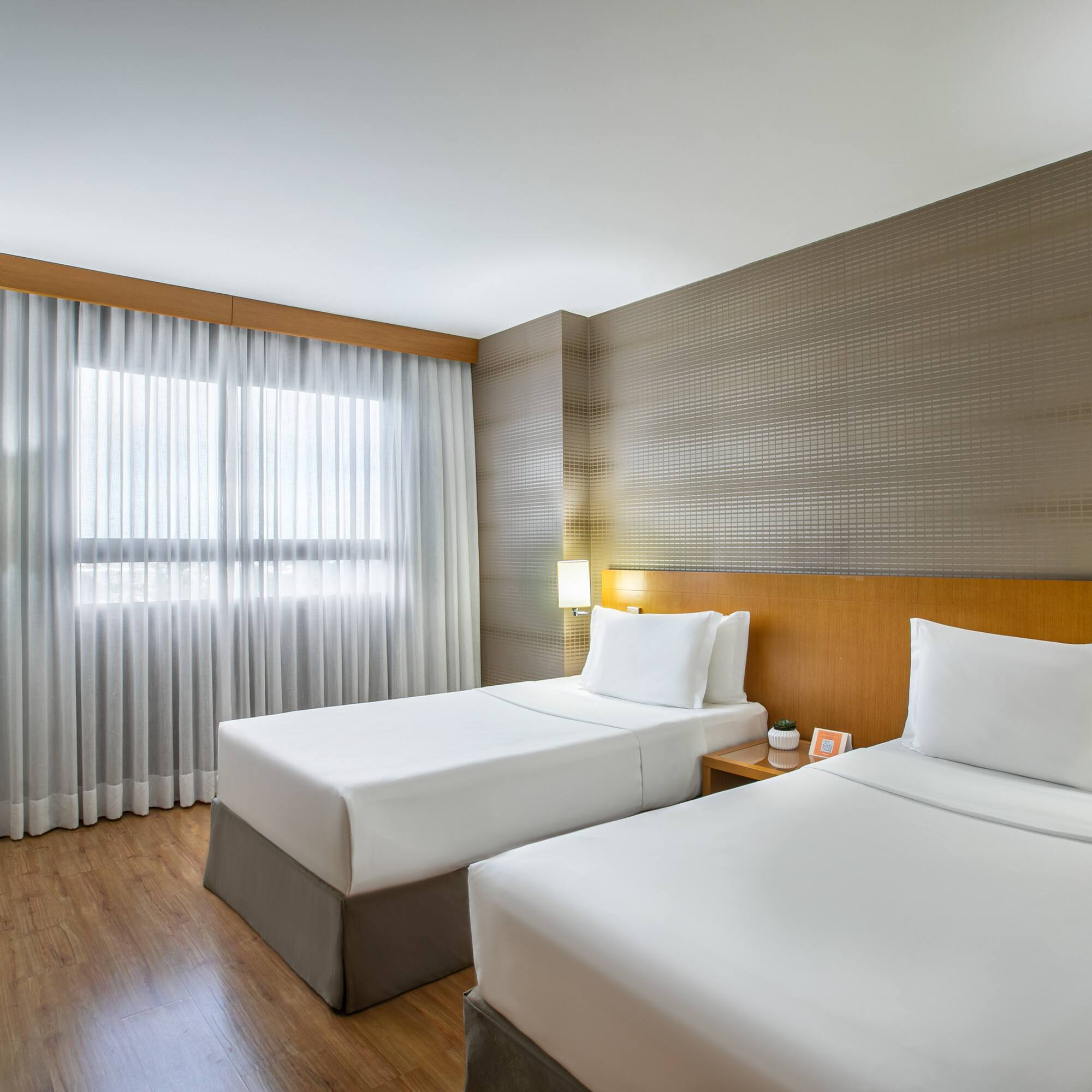 Modern hotel guest room with two twin beds, wood decor, and bright window.