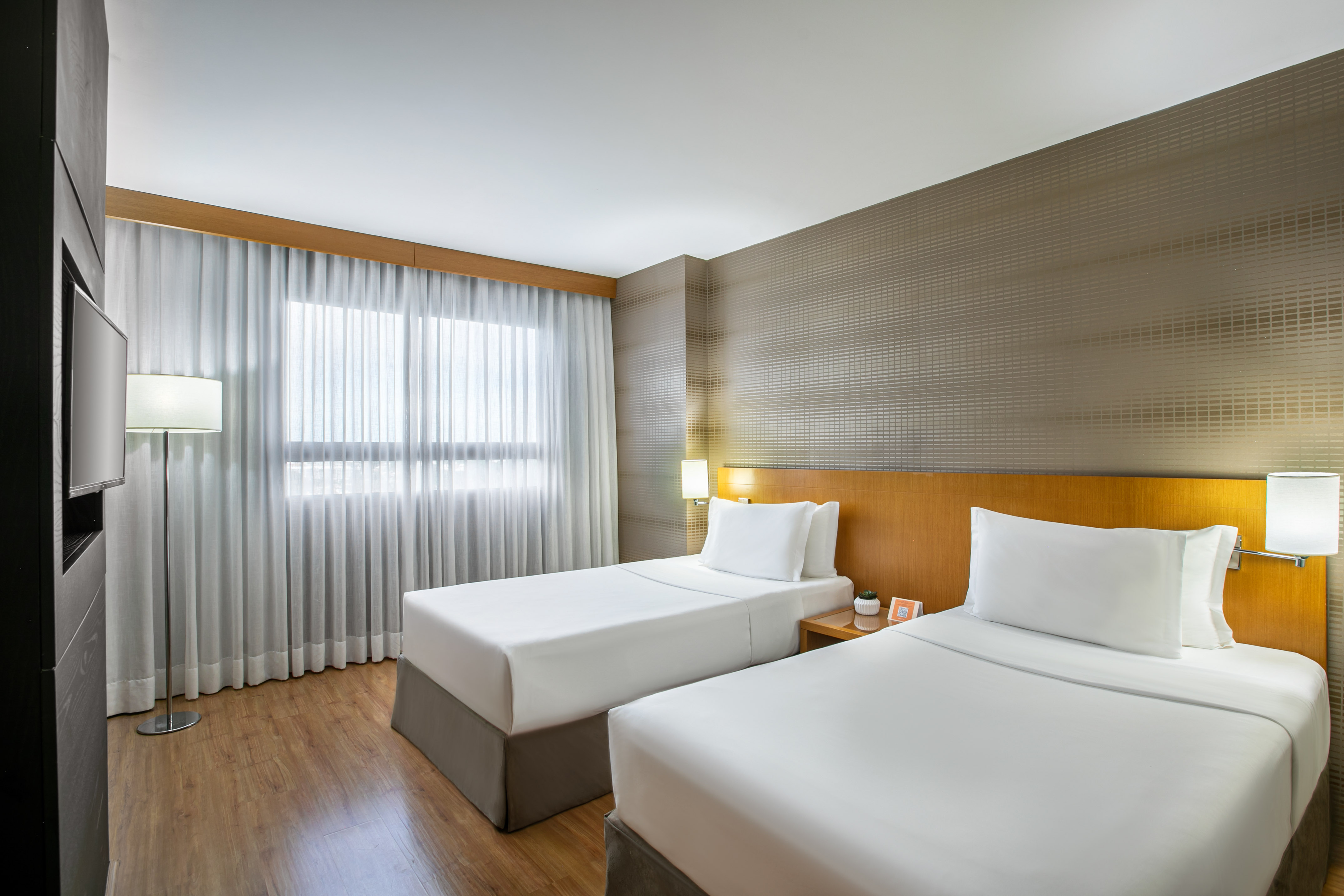 Modern hotel guest room with two twin beds, wood decor, and bright window.