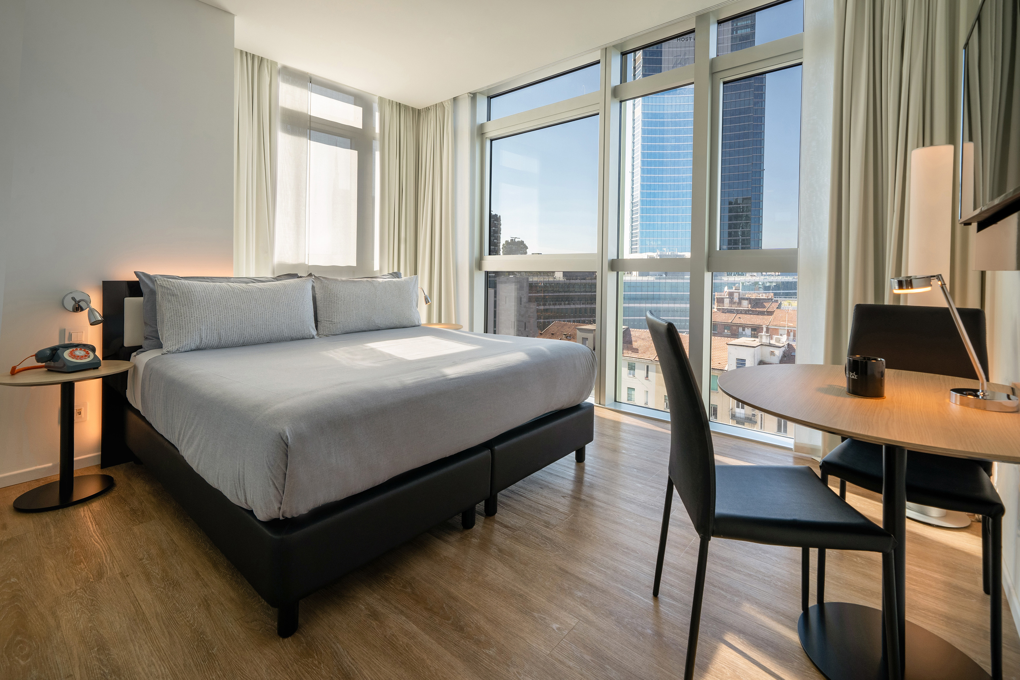 Modern hotel room with large bed, city view through big windows, and desk.