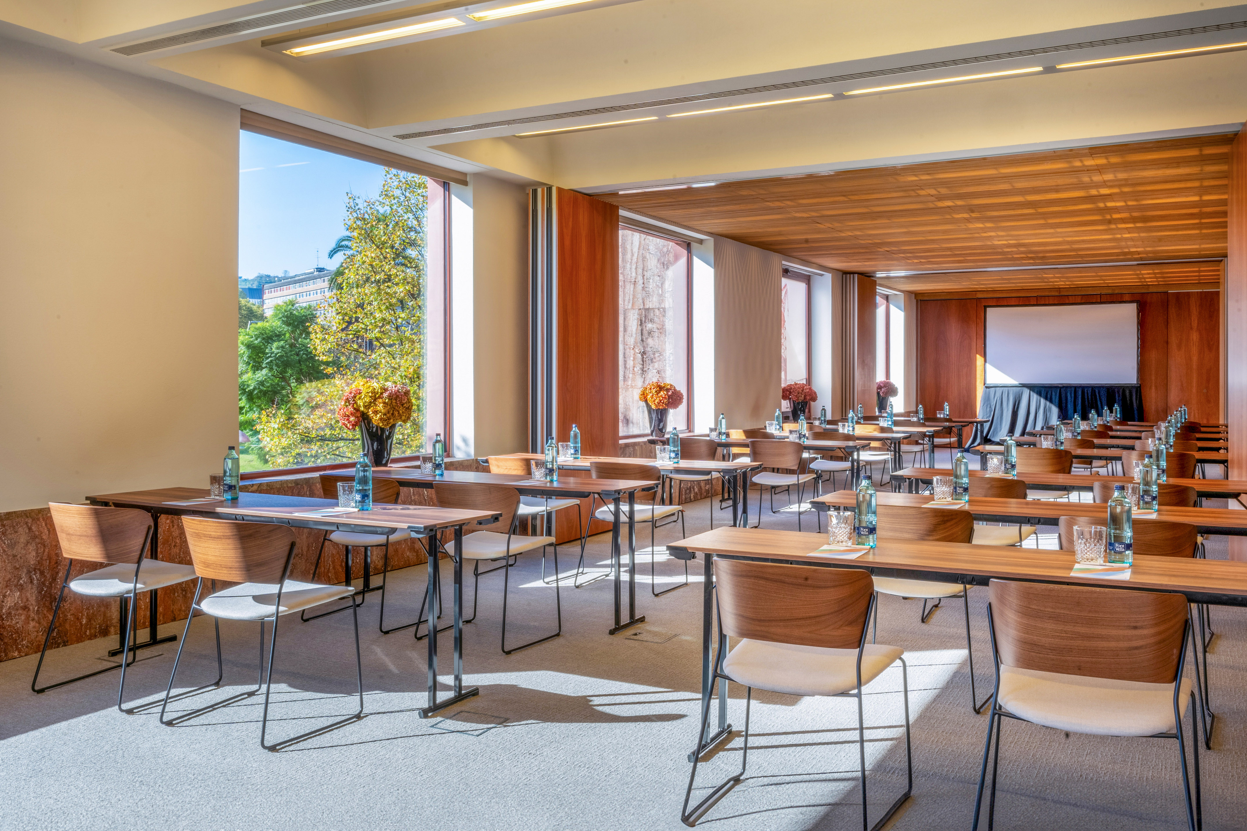 Well-lit conference room with tables, chairs, projection screen, and green outdoor view.