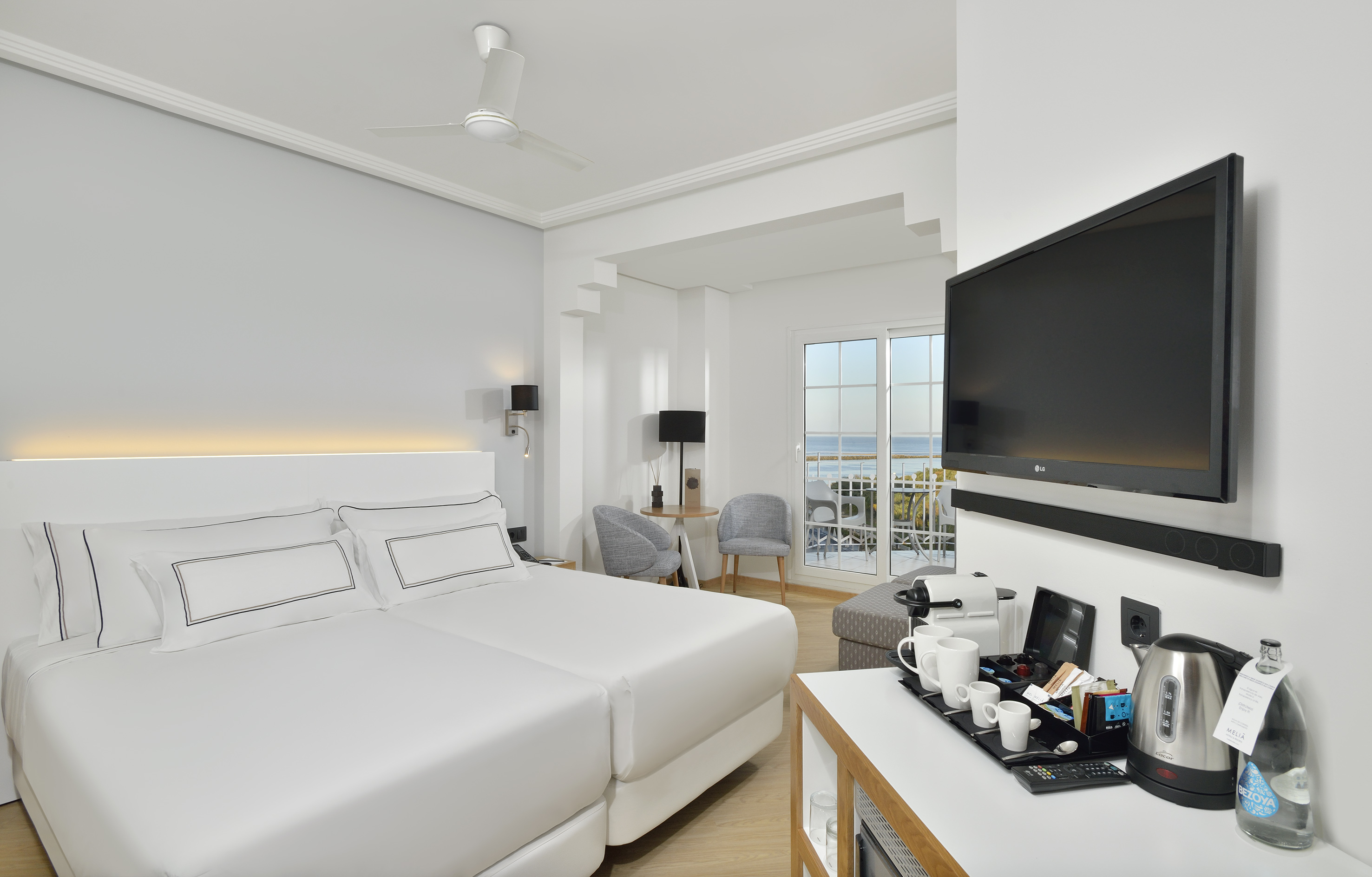 Modern hotel room with twin beds, ocean view balcony, TV, and coffee station.