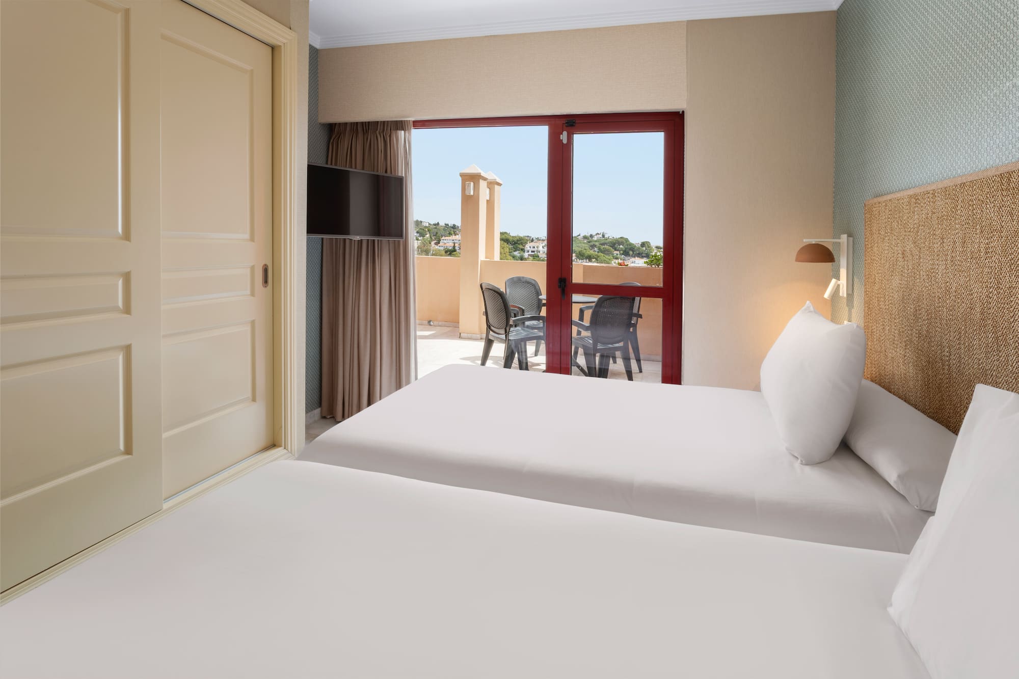 Bright twin hotel room with private balcony, outdoor furniture, and sunny view.