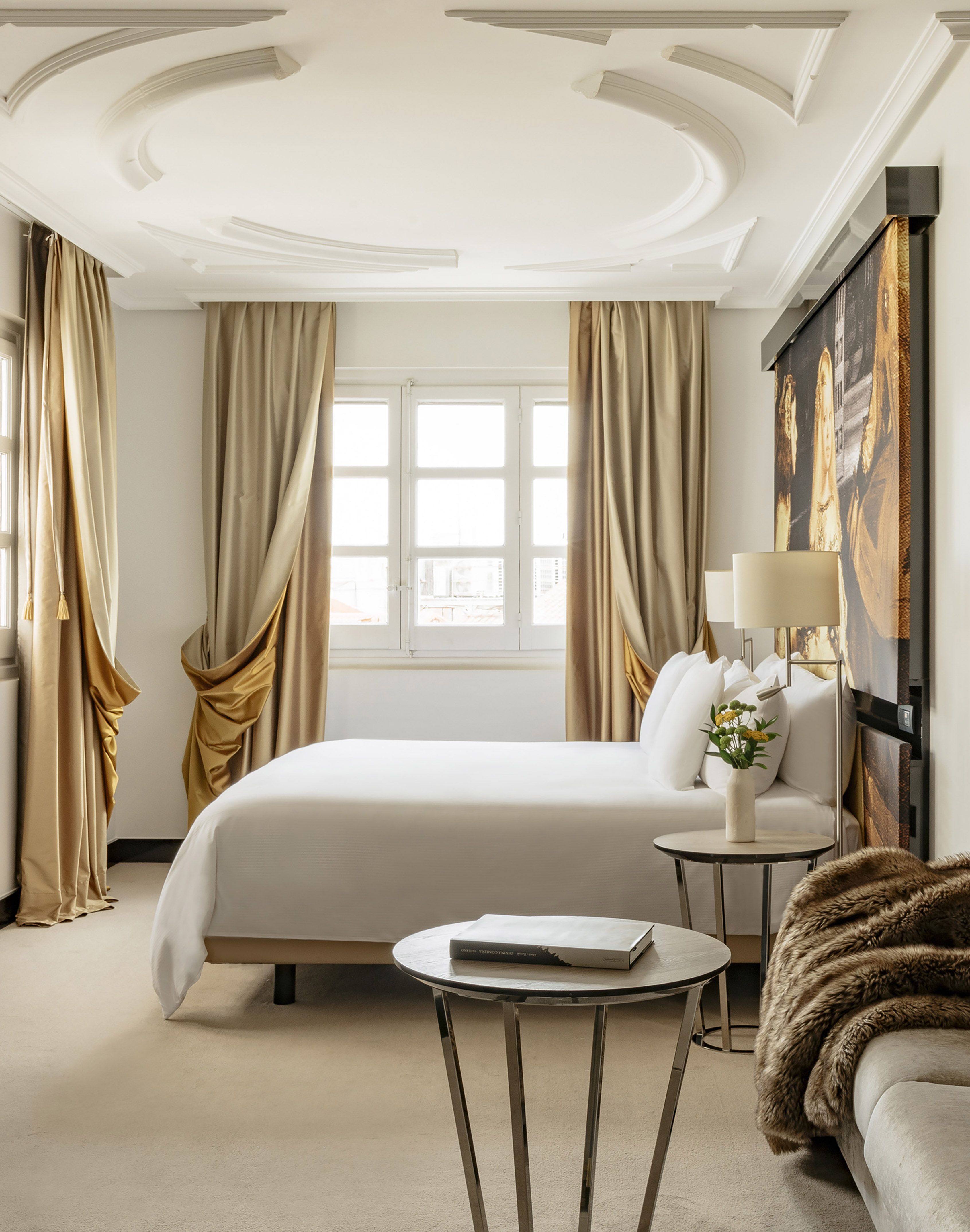 Luxury hotel bedroom with king bed, ornate ceiling, gold curtains, and wall art.