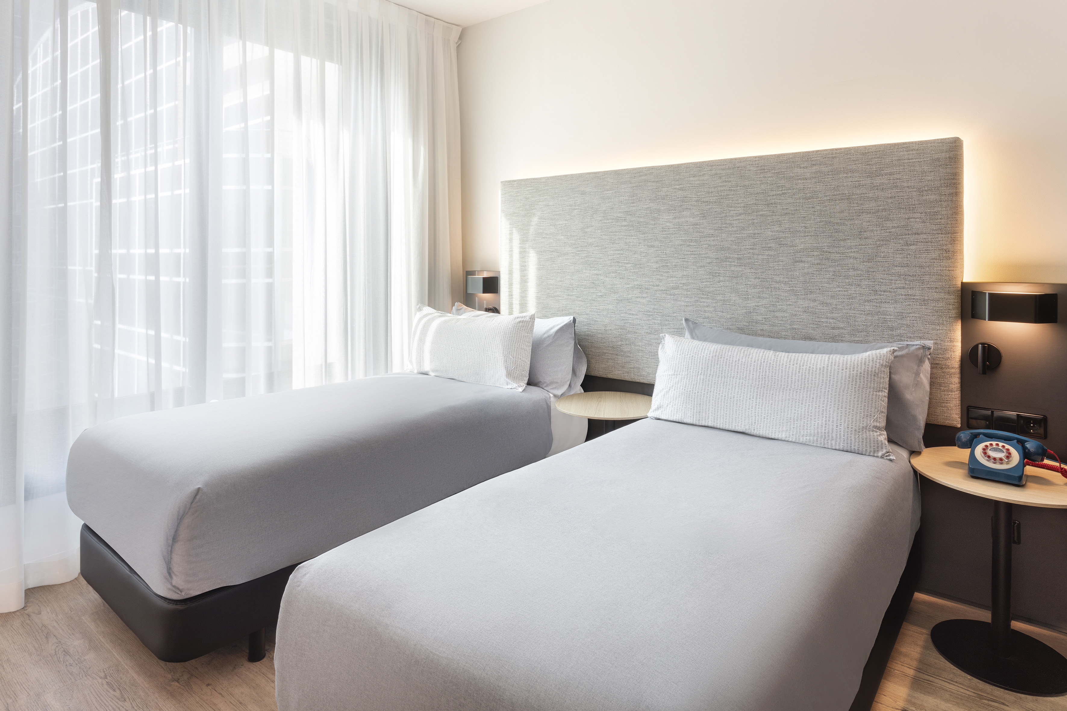 Modern hotel room with two single beds, grey bedding, and backlit headboard.
