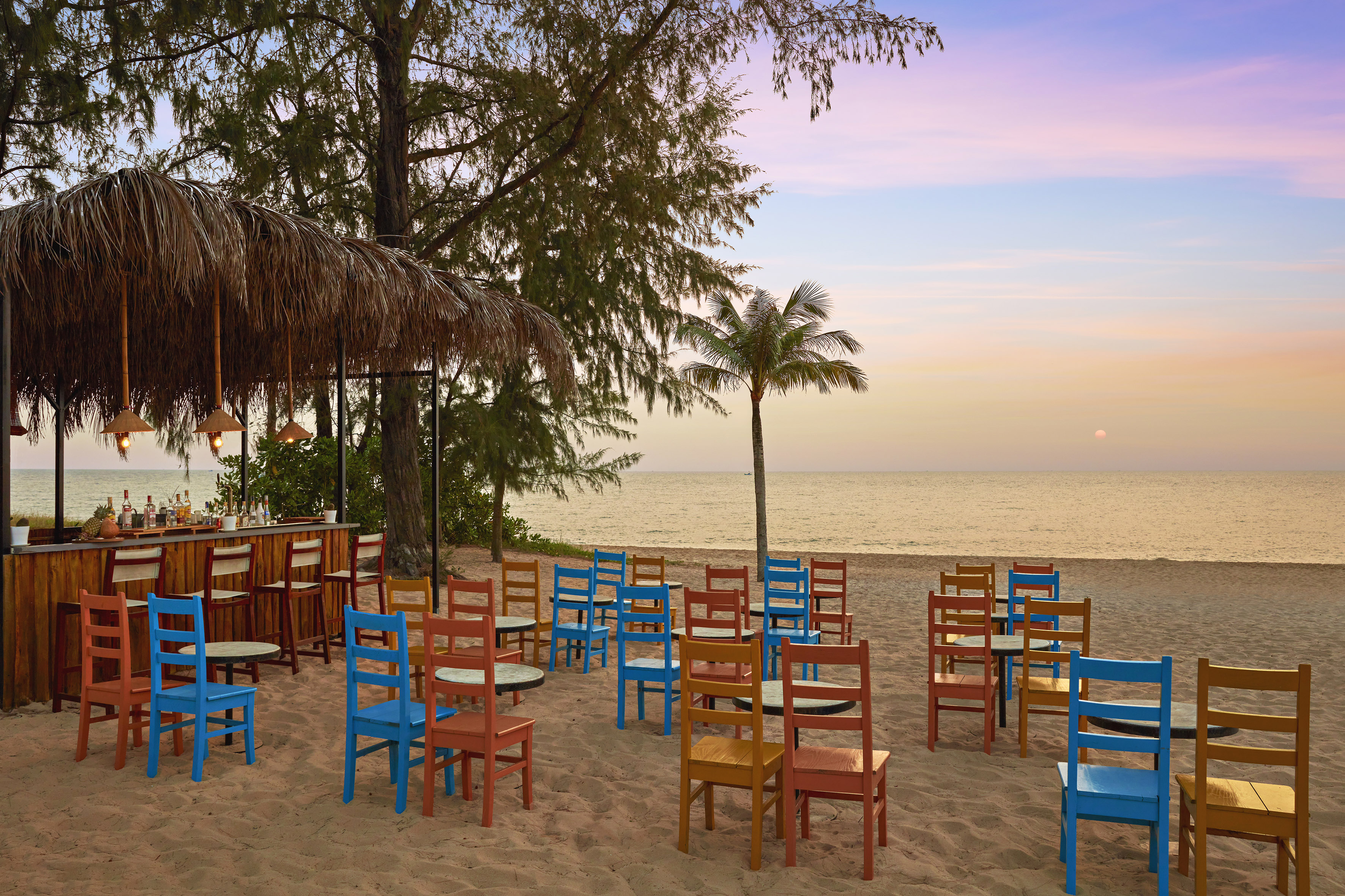Tropical beach bar with colorful chairs on sand, ocean sunset background.