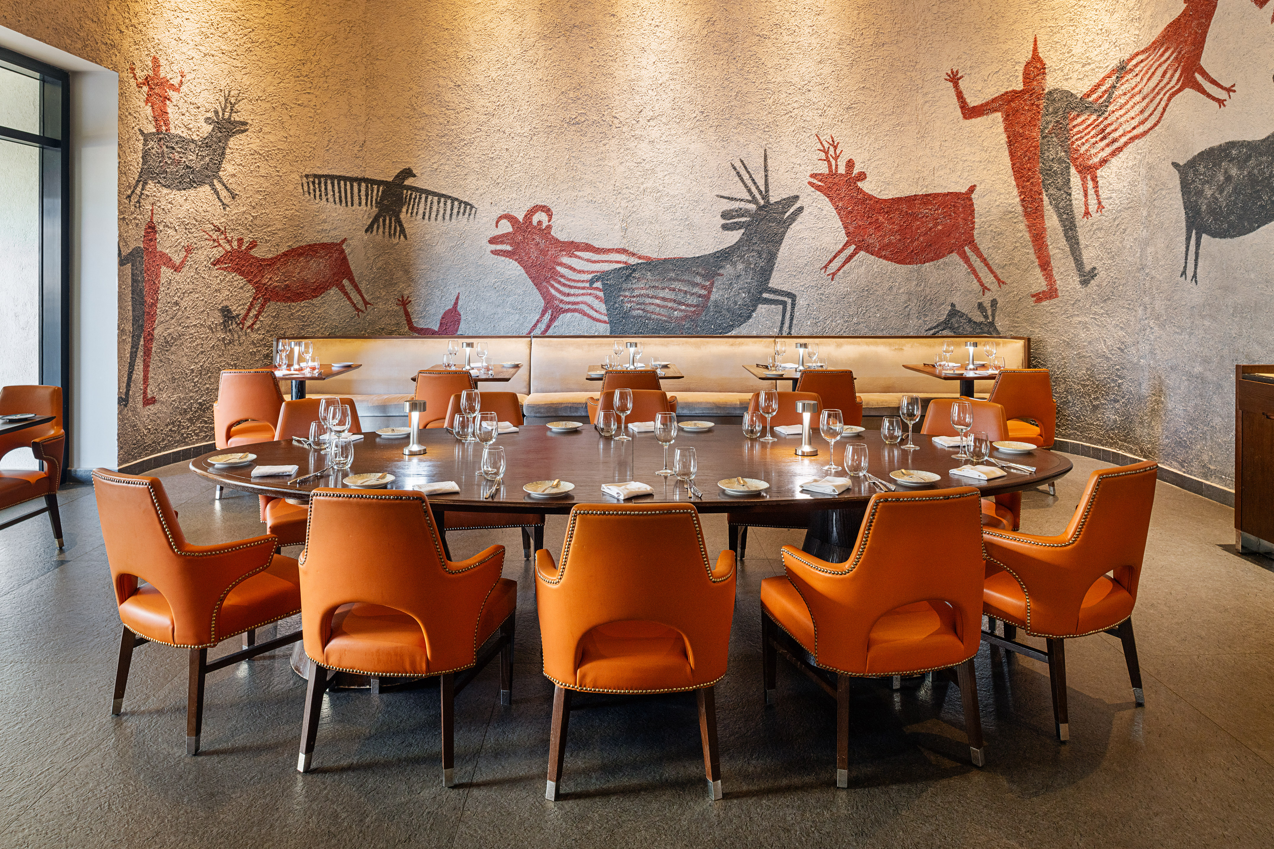 Oval dining table with orange leather chairs and cave painting wall mural.