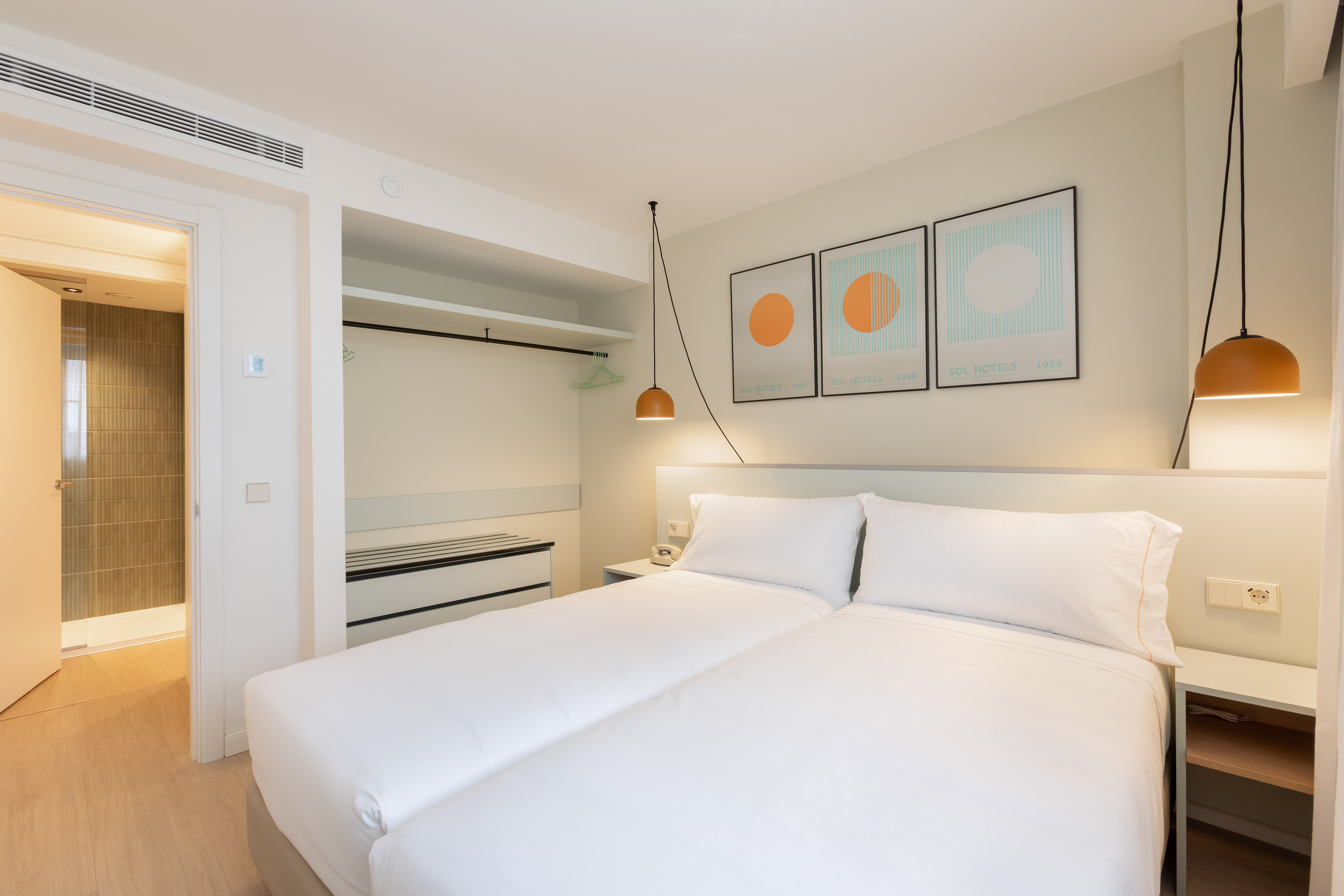 Modern hotel bedroom with twin beds, abstract art, and open wardrobe.