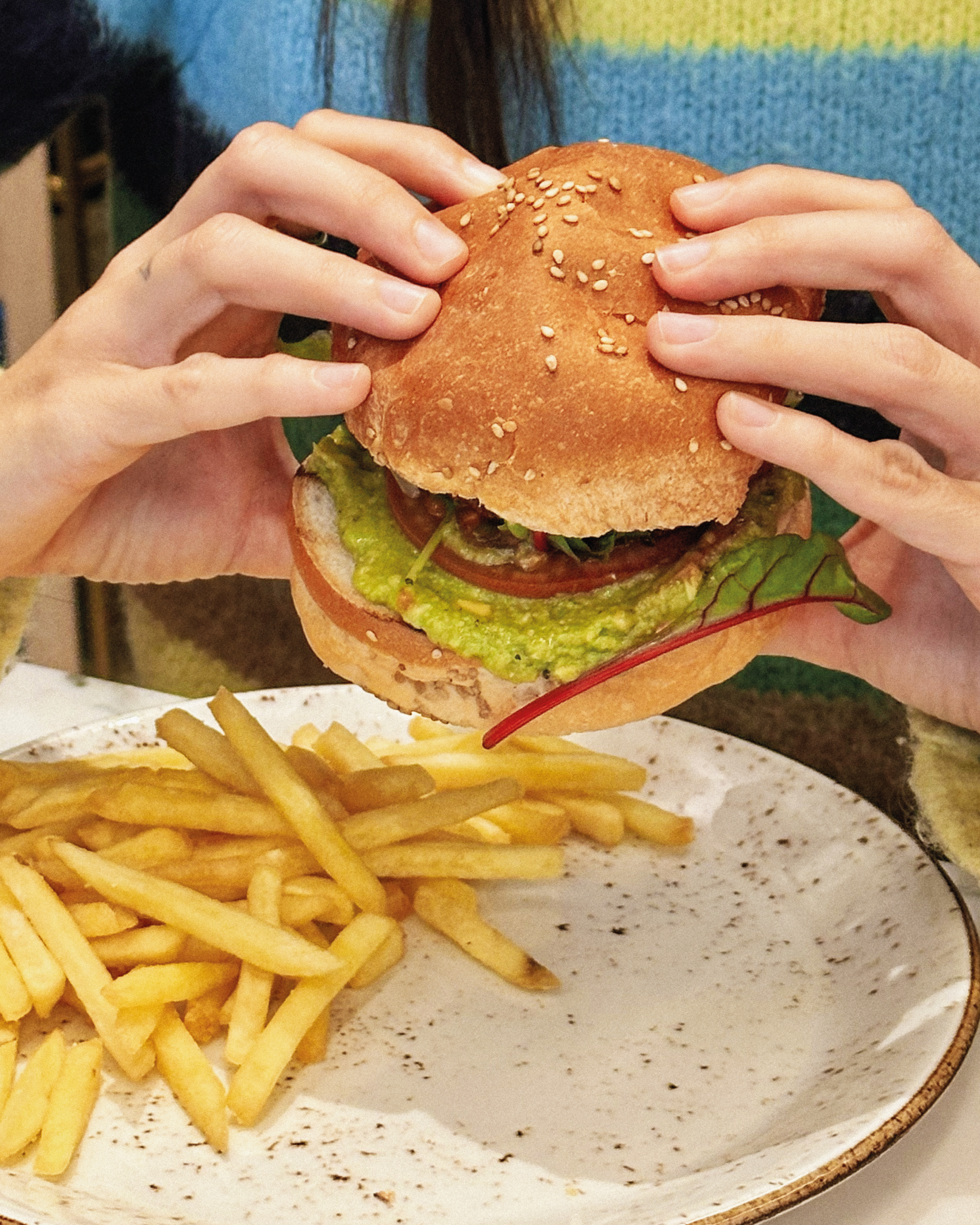 Hands holding a guacamole burger with fries on a plate.